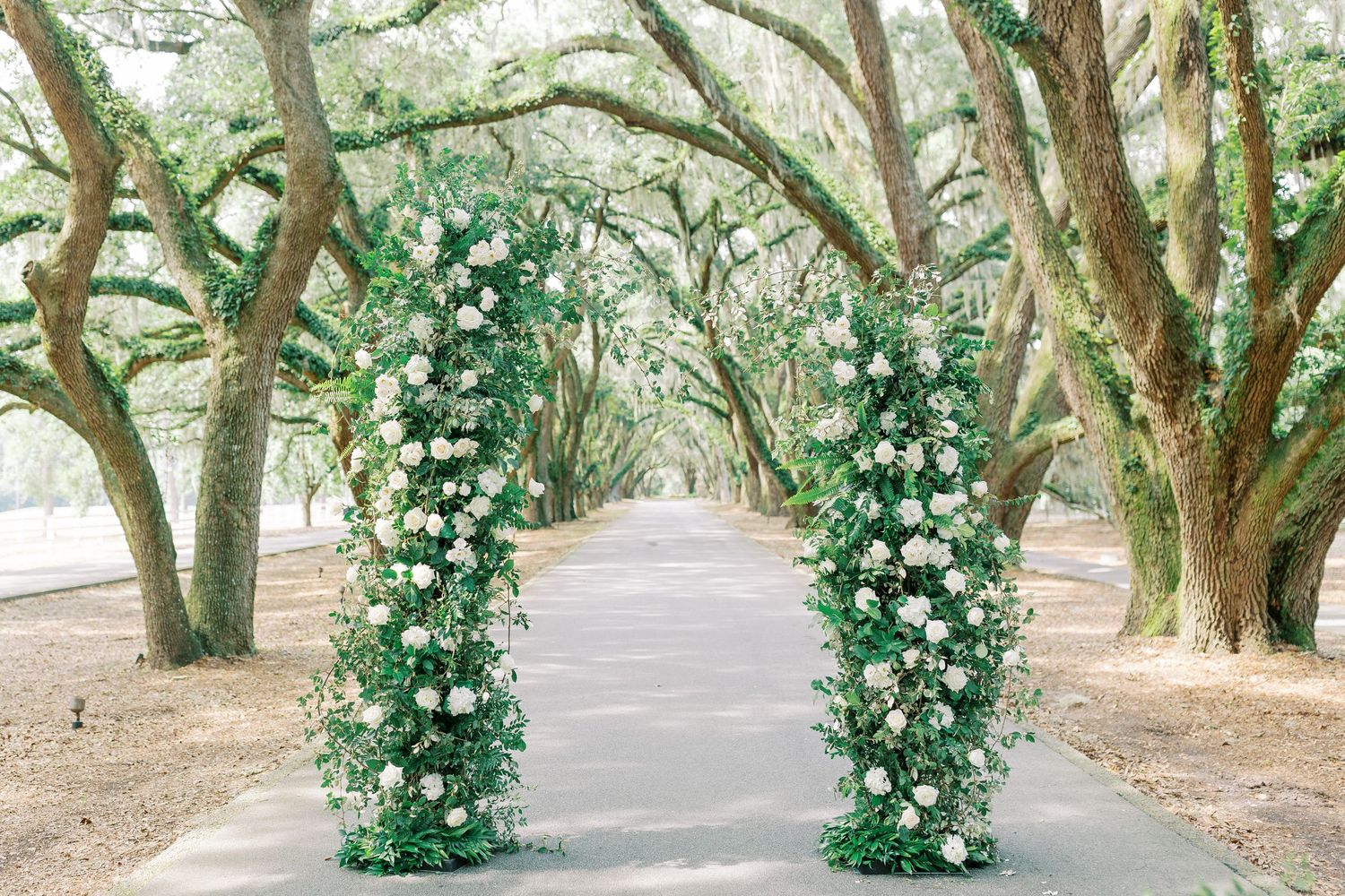 Floral wedding arch with white roses and greenery creates a tunnel effect on a tree-lined pathway.