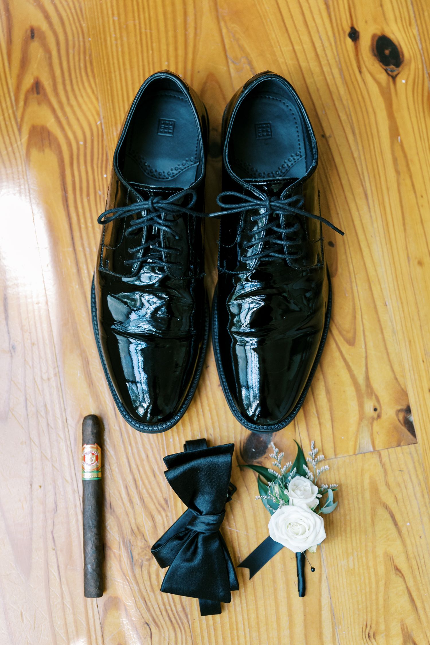 Black patent leather dress shoes arranged with a cigar, black bow tie, and white rose boutonniere on wooden floor.