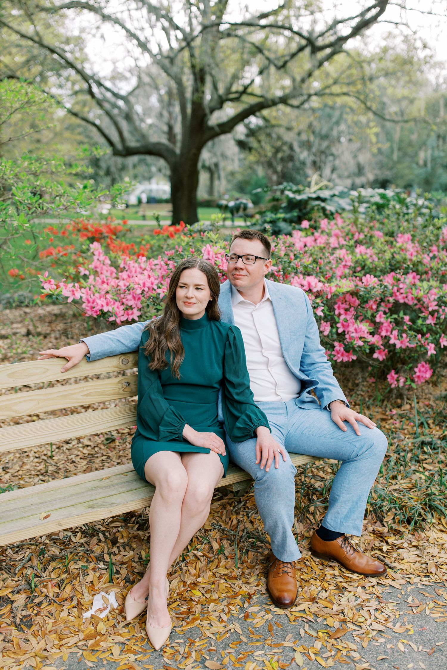 A couple sits on a wooden bench surrounded by blooming pink azalea bushes and trees in a garden setting.