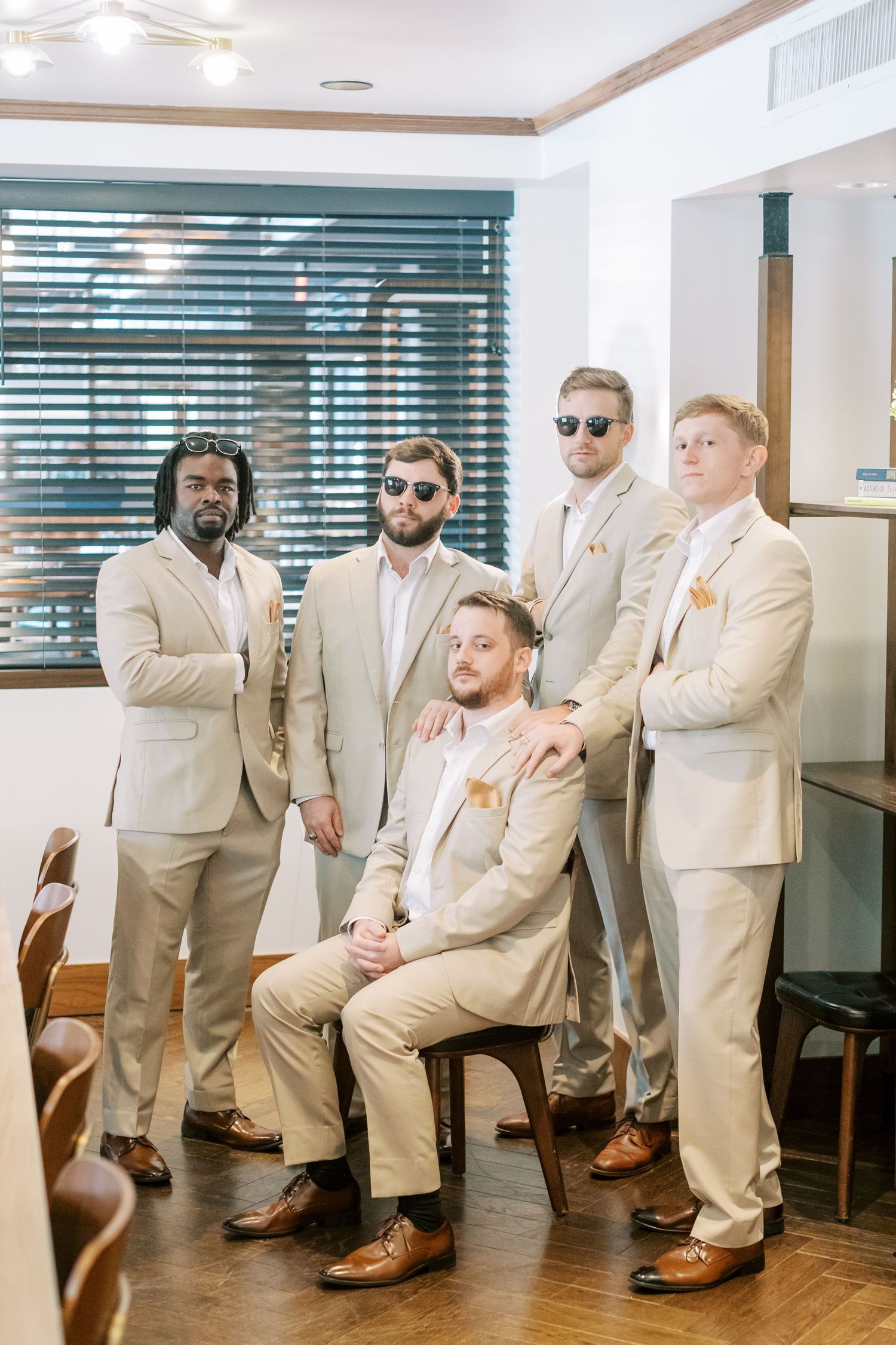 Groomsmen in tan suits strike fun poses while lined up against a white wall.
