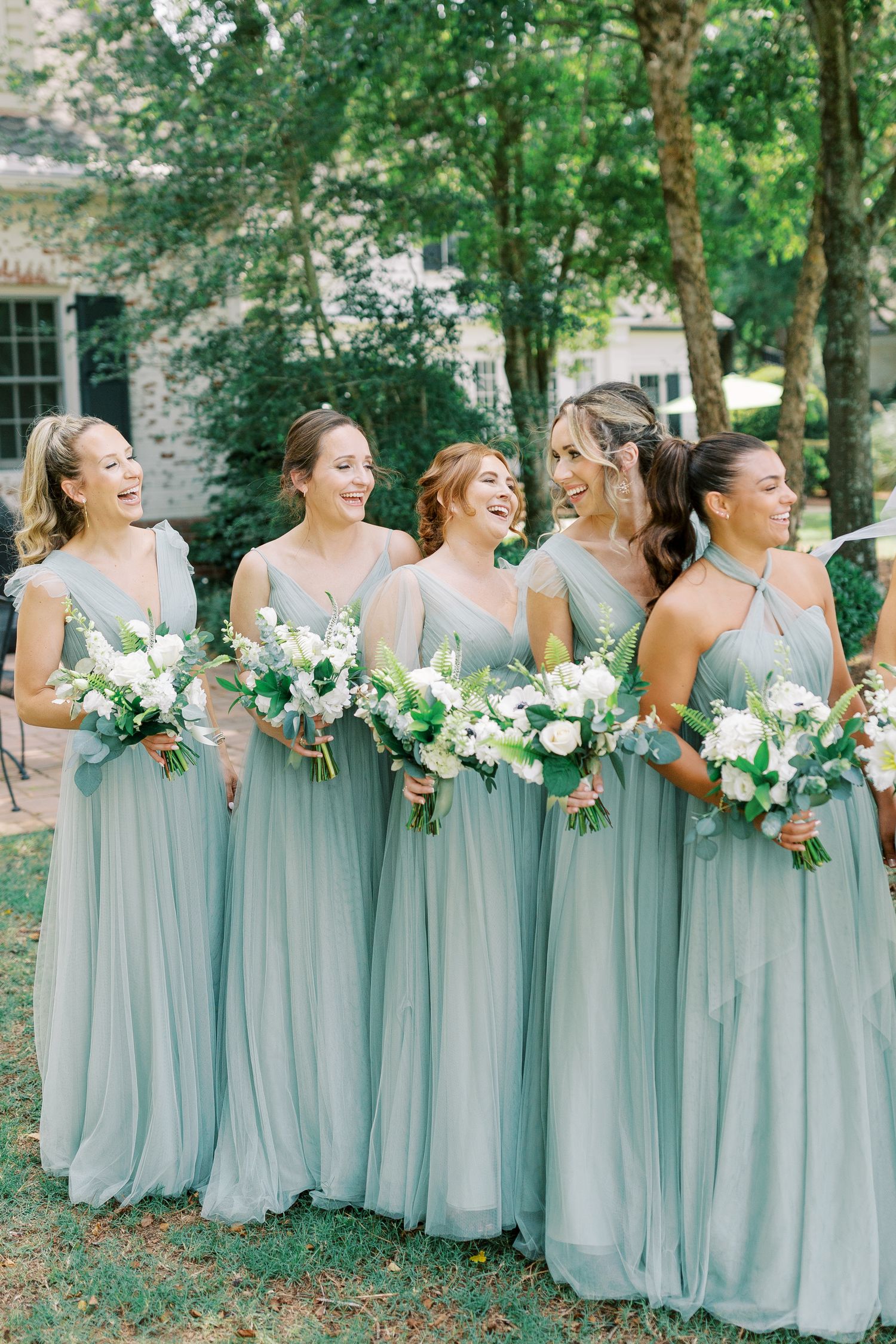 Bridesmaids in flowing sage green dresses holding white and green bouquets laugh together in an outdoor garden setting.