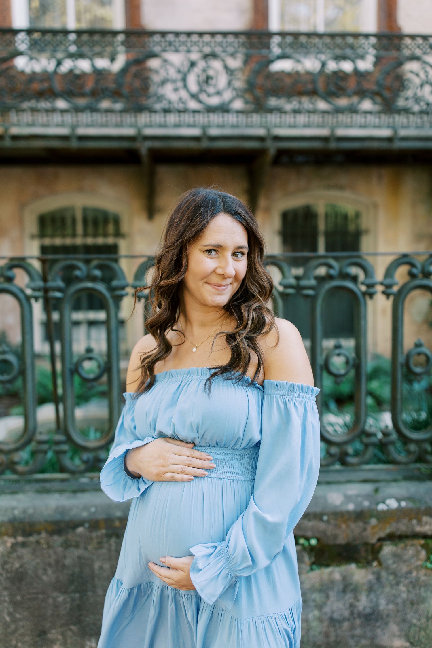 A woman in a light blue off-shoulder maternity dress poses by vintage wrought iron railings.