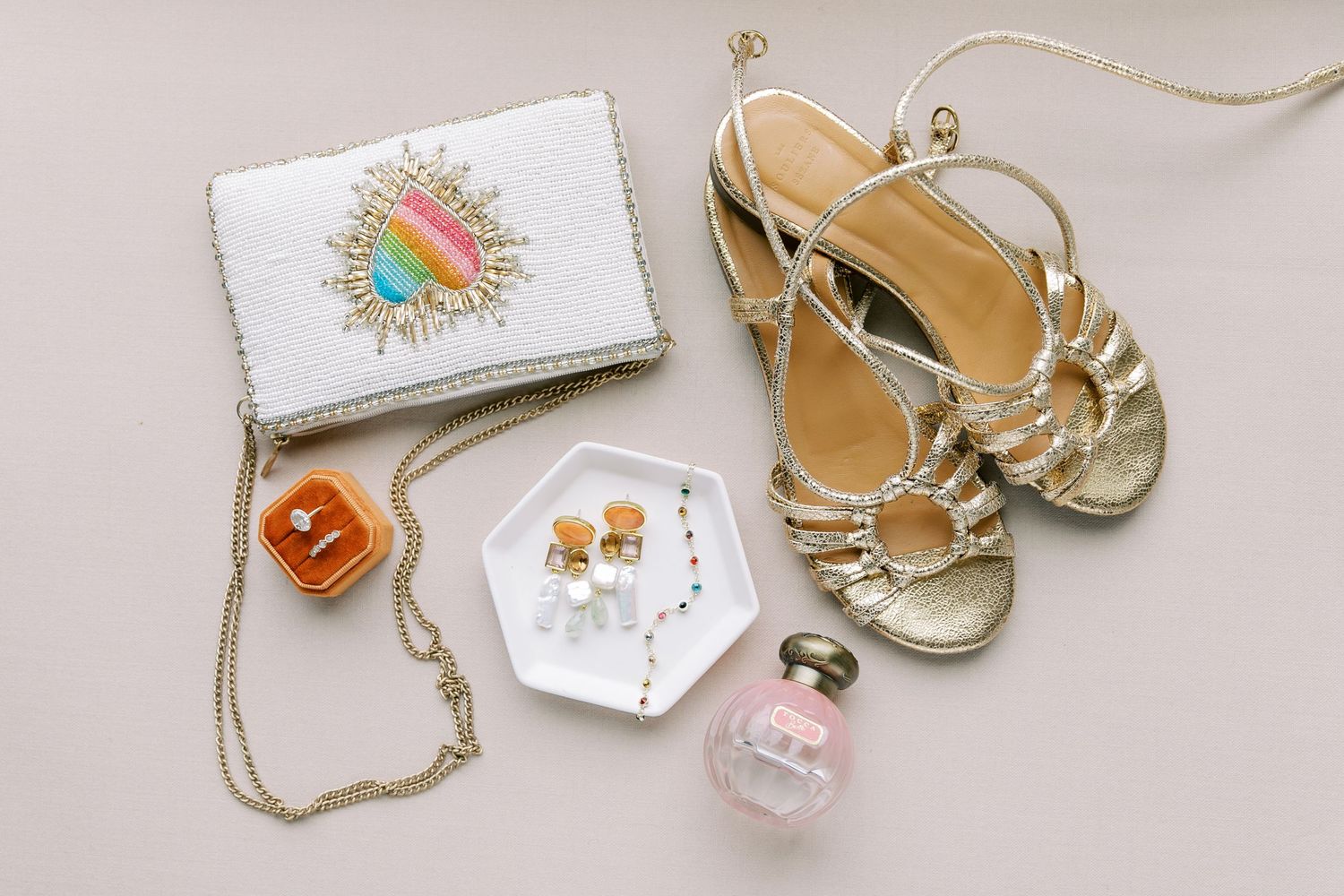 Elegant bridal accessories including gold sandals, perfume bottle, white handbag, and delicate jewelry laid out on beige surface.