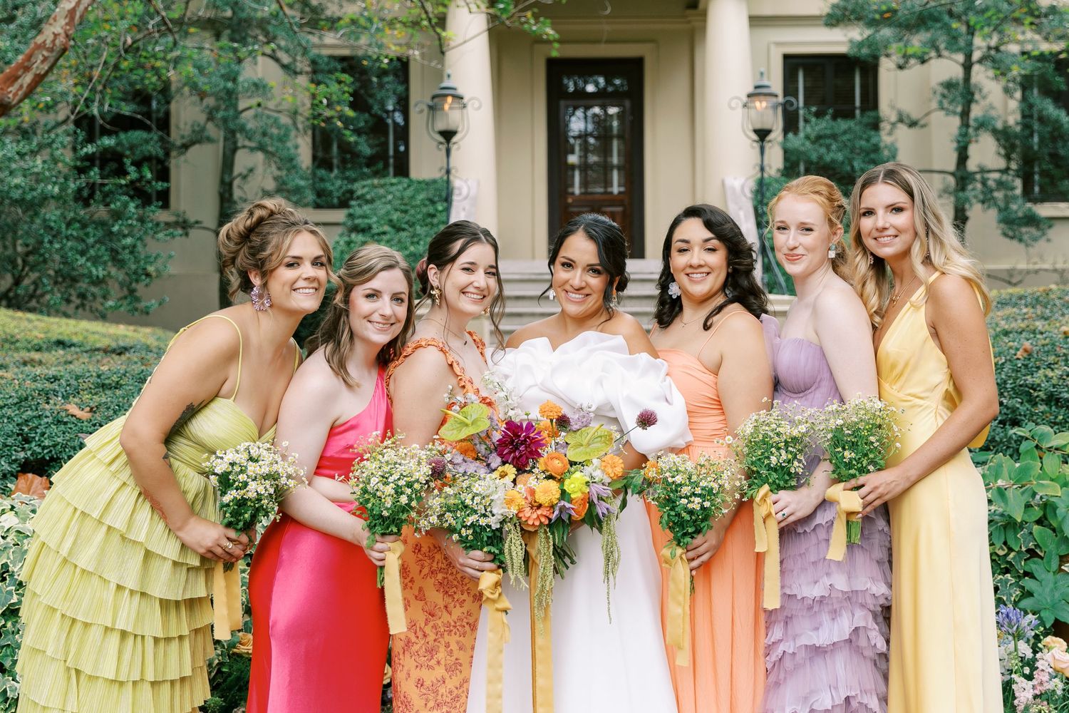 Bridesmaids wearing colorful pastel dresses pose together with bouquets in a garden setting.