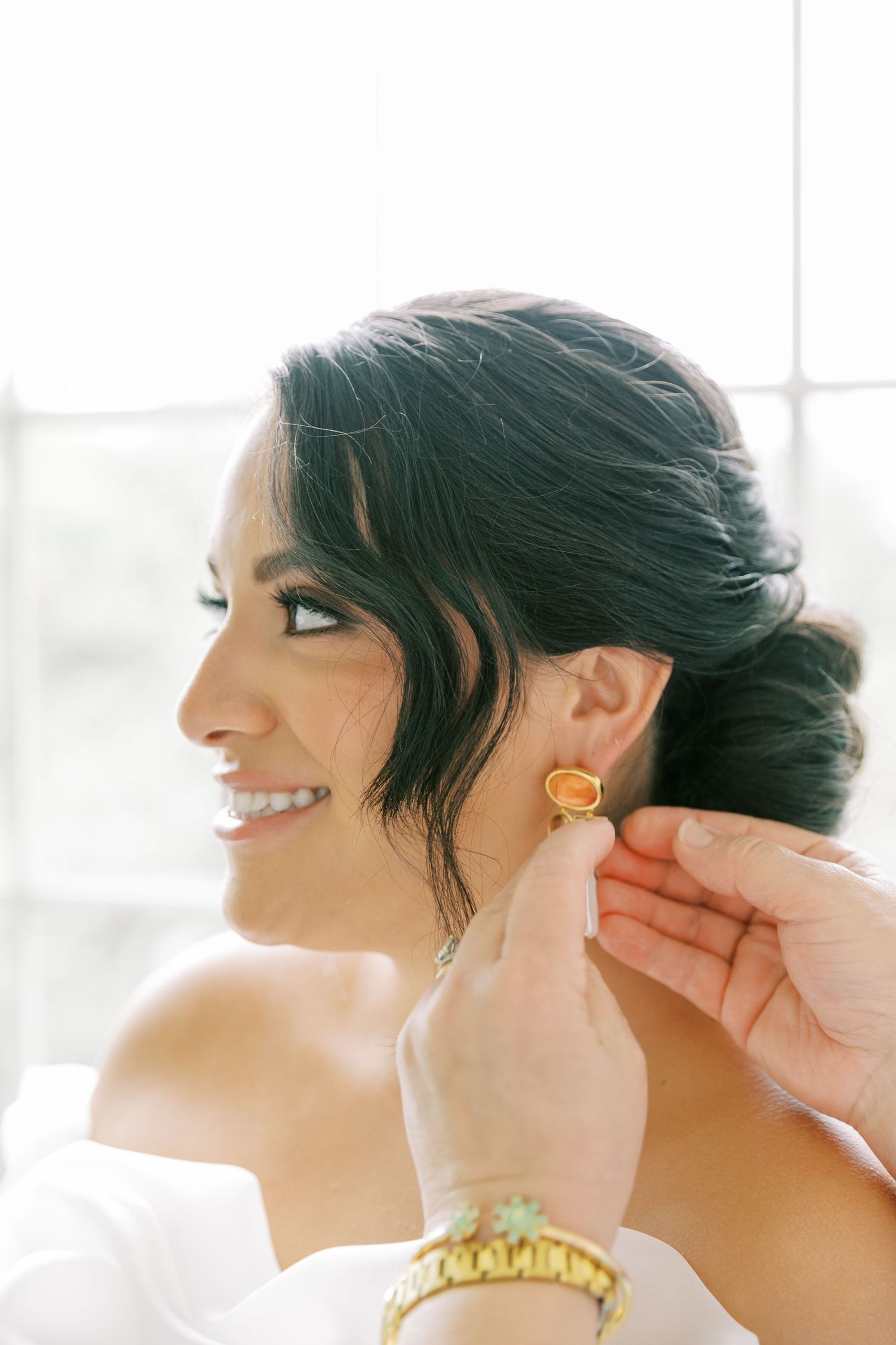 Smiling person getting ready on their wedding day as someone helps put on gold earrings.
