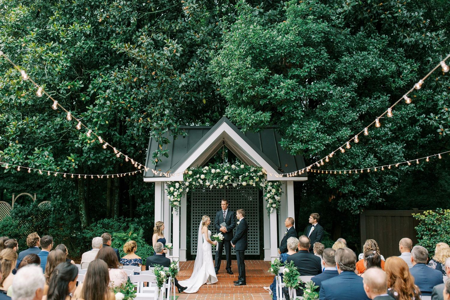 Outdoor wedding ceremony at a small chapel in the woods with string lights and guests seated on green folding chairs in Norcross, GA