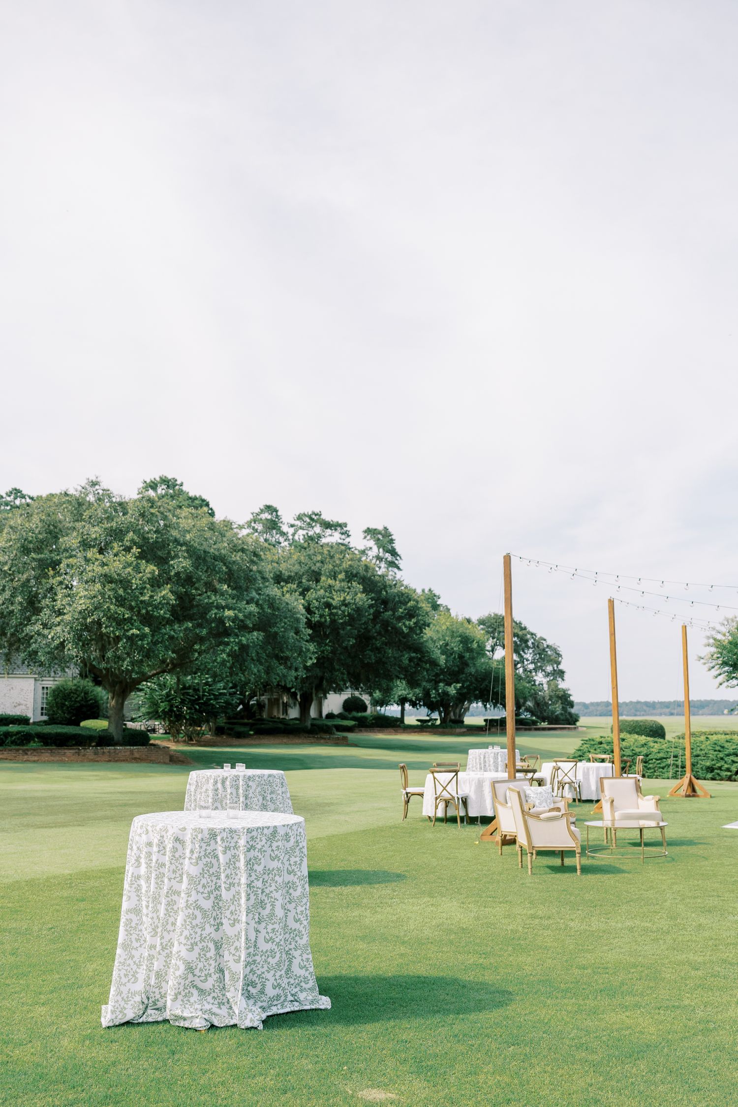 Outdoor wedding reception setup on a manicured lawn with string lights and a white patterned linen cocktail table.