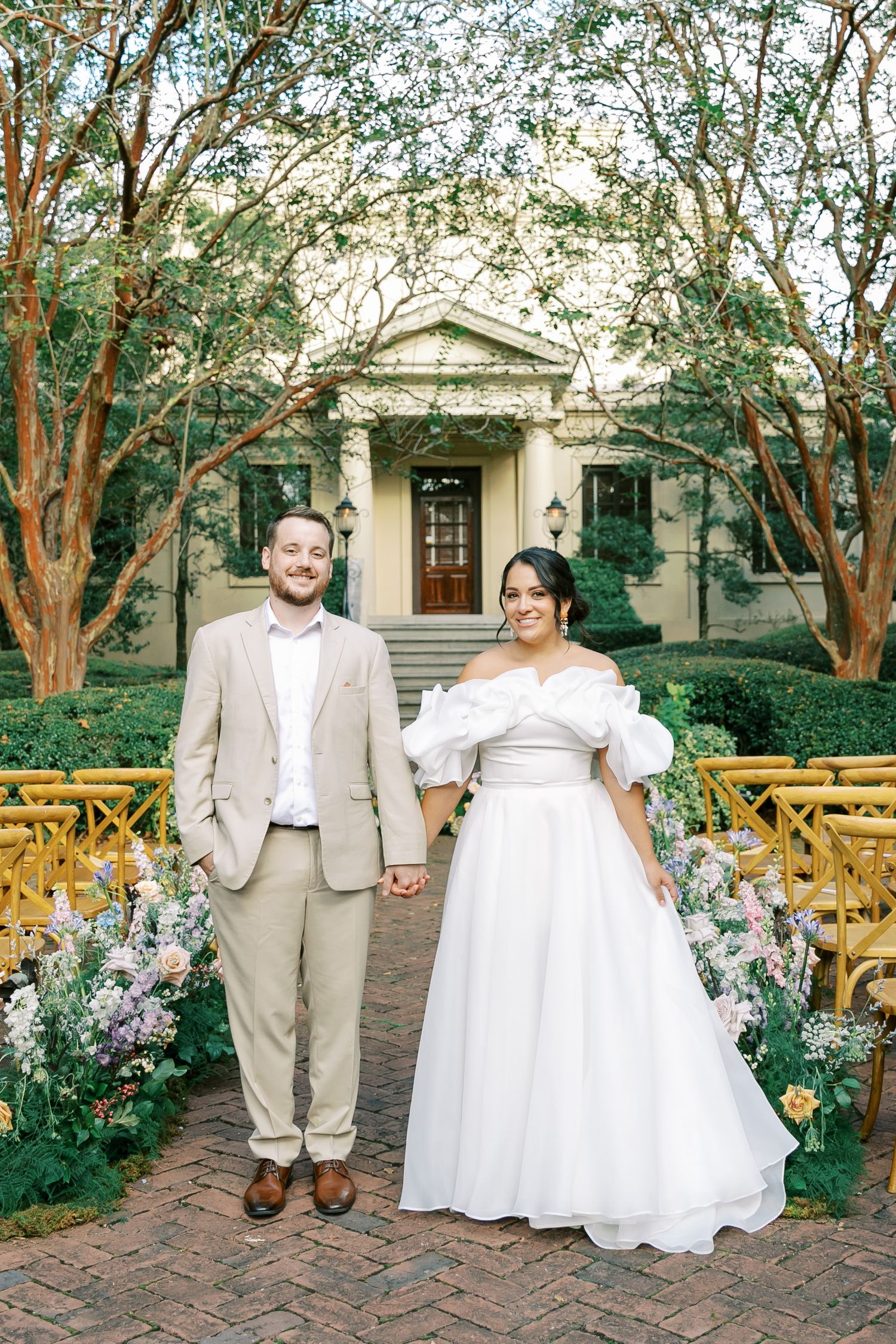 A couple poses for wedding photos on a brick pathway surrounded by lush garden landscaping.