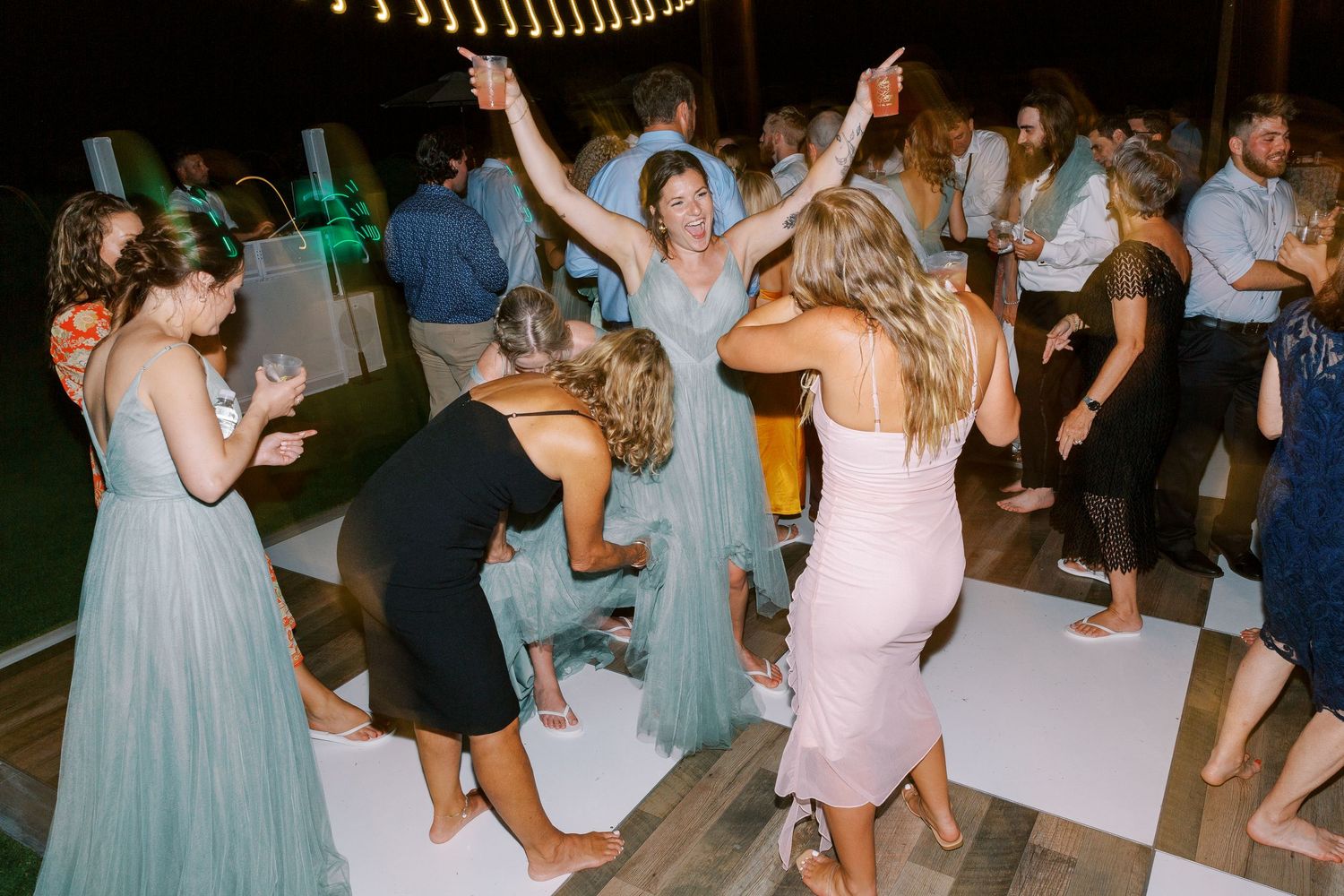 Wedding guests dance energetically on a white dance floor under outdoor lighting during an evening reception.