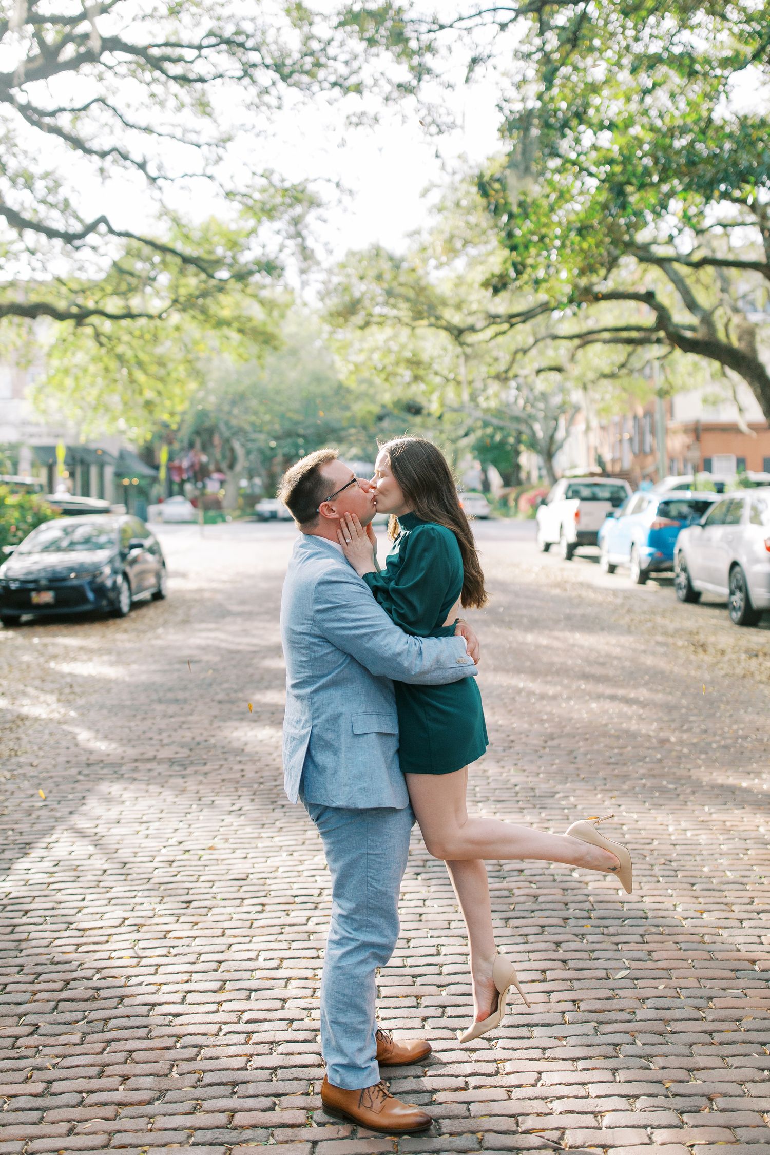 A couple shares a romantic embrace on a brick-paved street lined with parked cars.