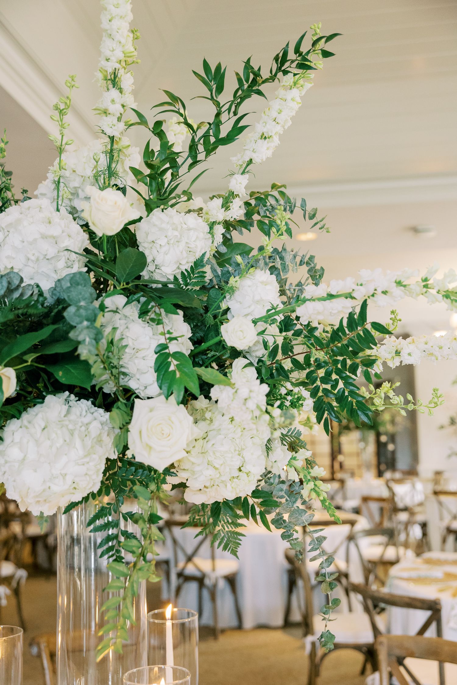 Elegant white floral centerpiece with hydrangeas and greenery in a tall glass vase at an upscale wedding reception.