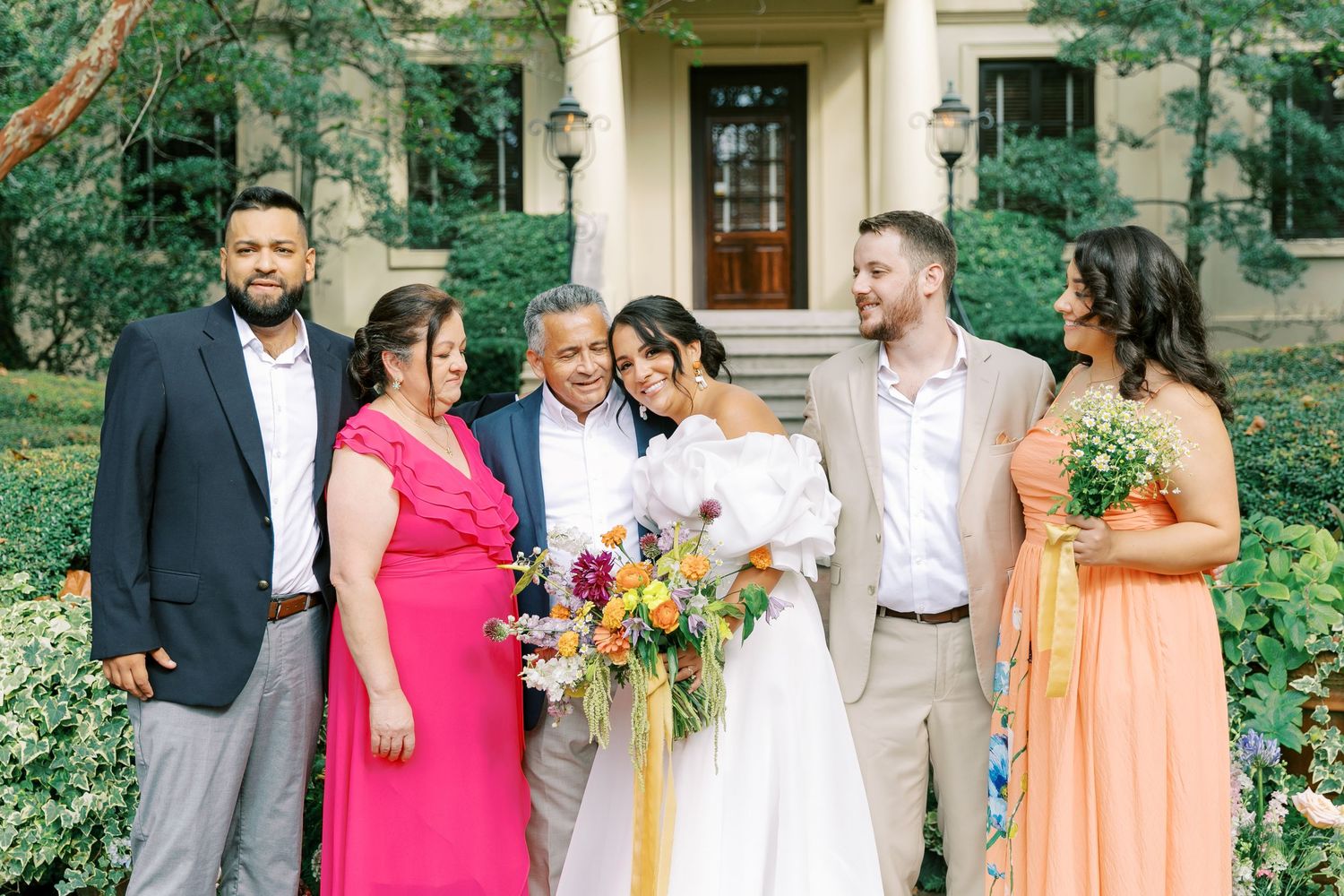 Wedding party poses for outdoor photos with bridesmaids in pink and orange dresses.