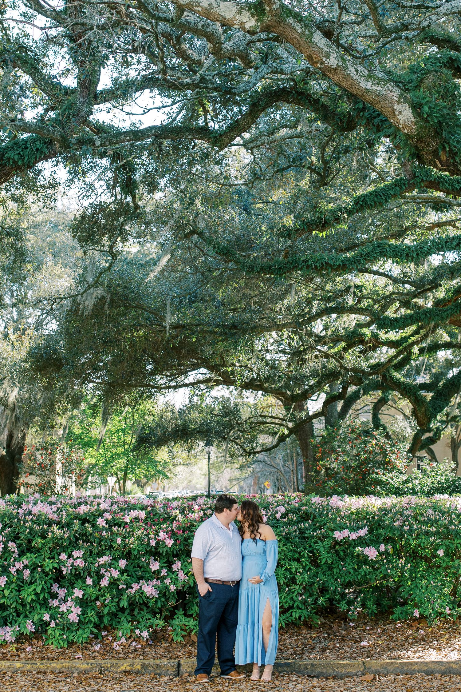 A romantic photoshoot under large oak trees with pink flowers blooming in the background.