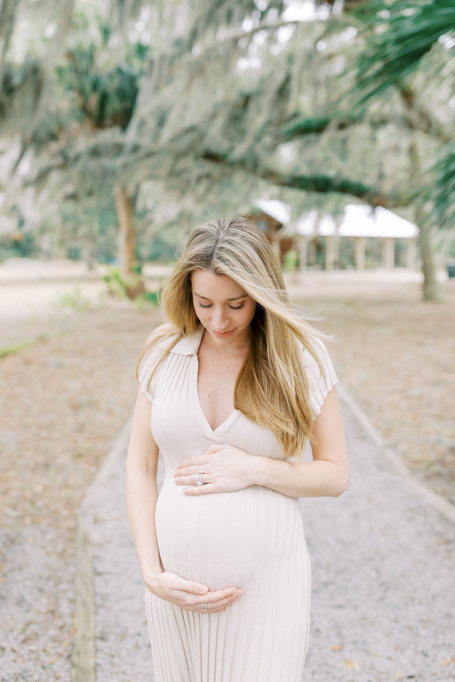 A pregnant woman in a flowing cream dress stands on a path beneath Spanish moss-draped trees in a serene outdoor setting.