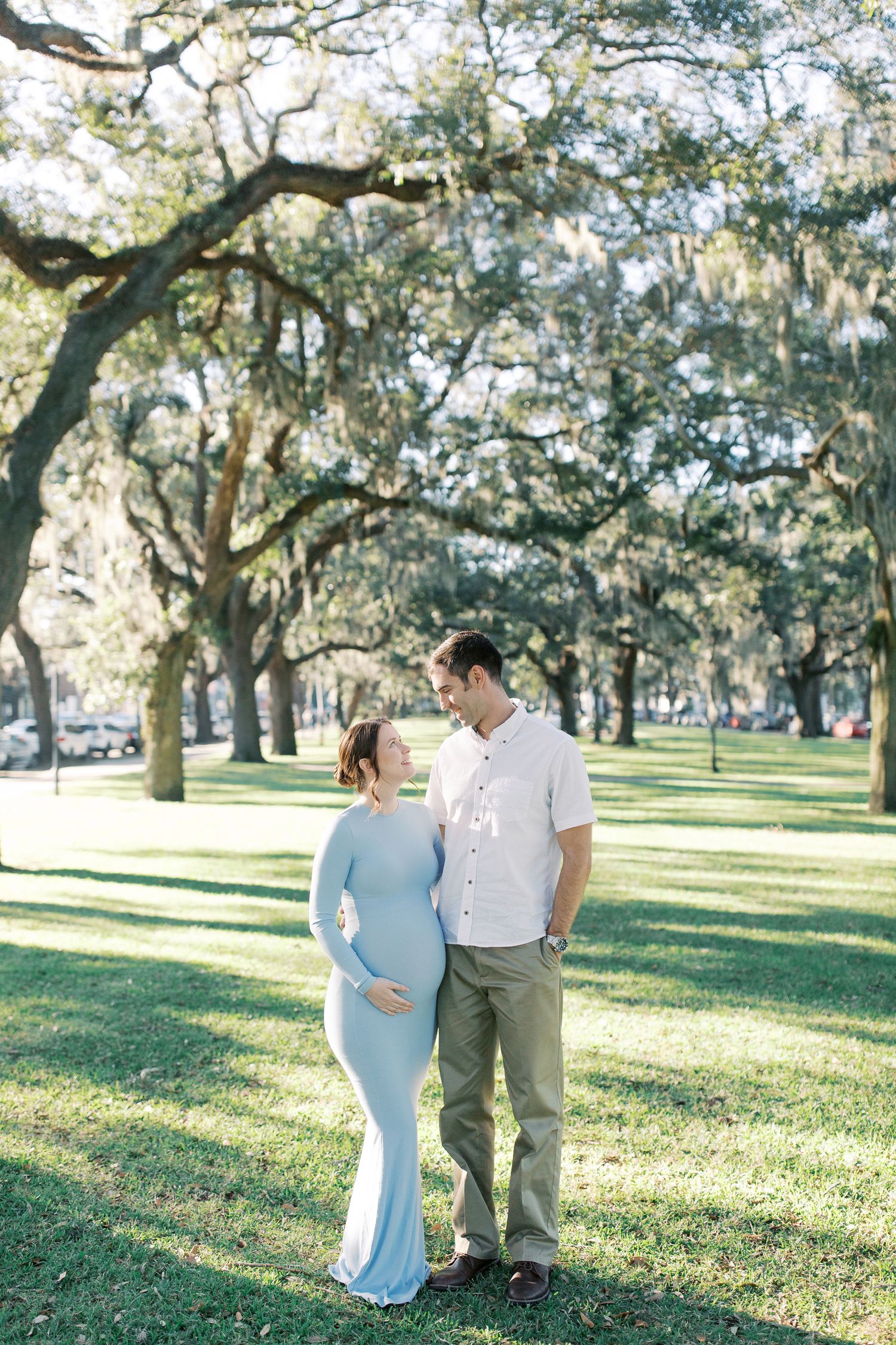 A couple stands together under Spanish moss-draped oak trees in a sunlit park setting during a maternity photo session.