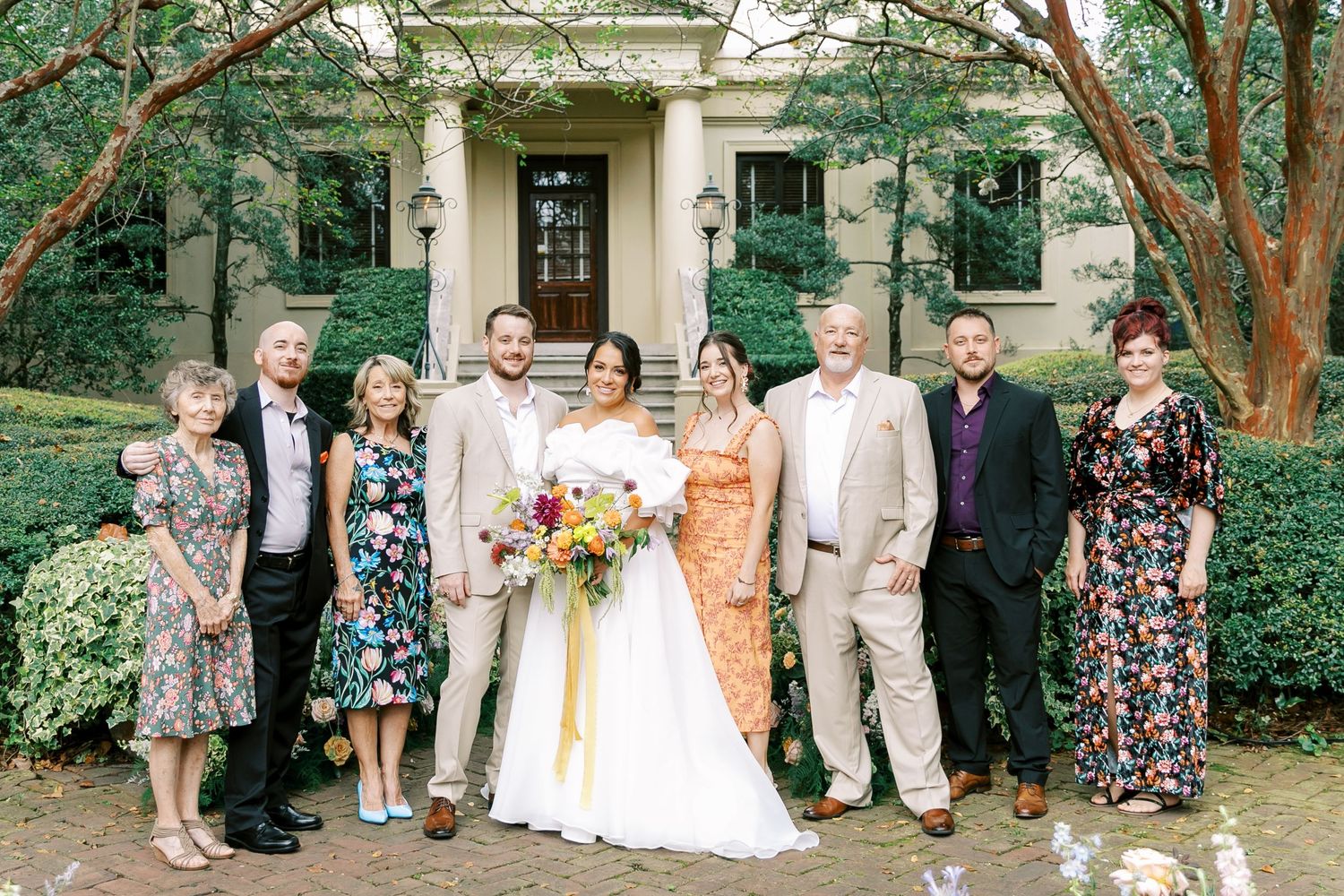 Wedding party group photo in front of historic building with floral landscaping.