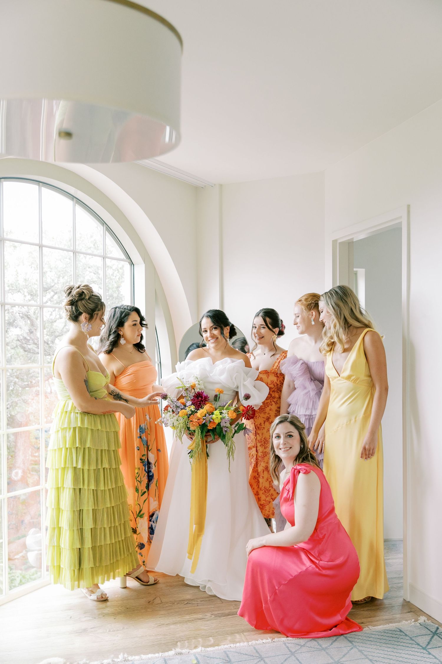 A group in colorful formal dresses including yellow, coral and pink gather by a large arched window for a special occasion.