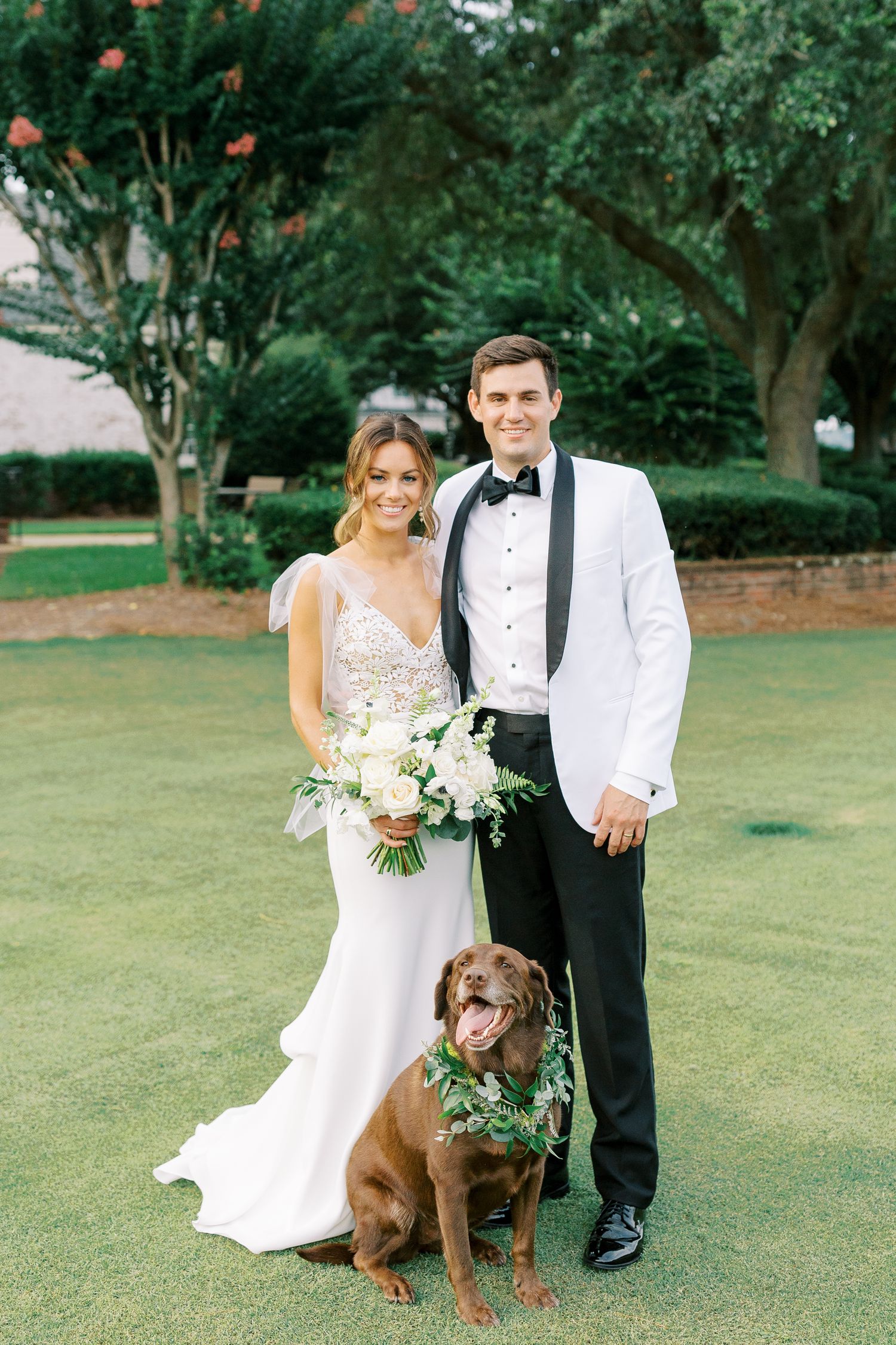 Wedding couple poses on green lawn with brown dog wearing leafy collar and white floral bouquet.