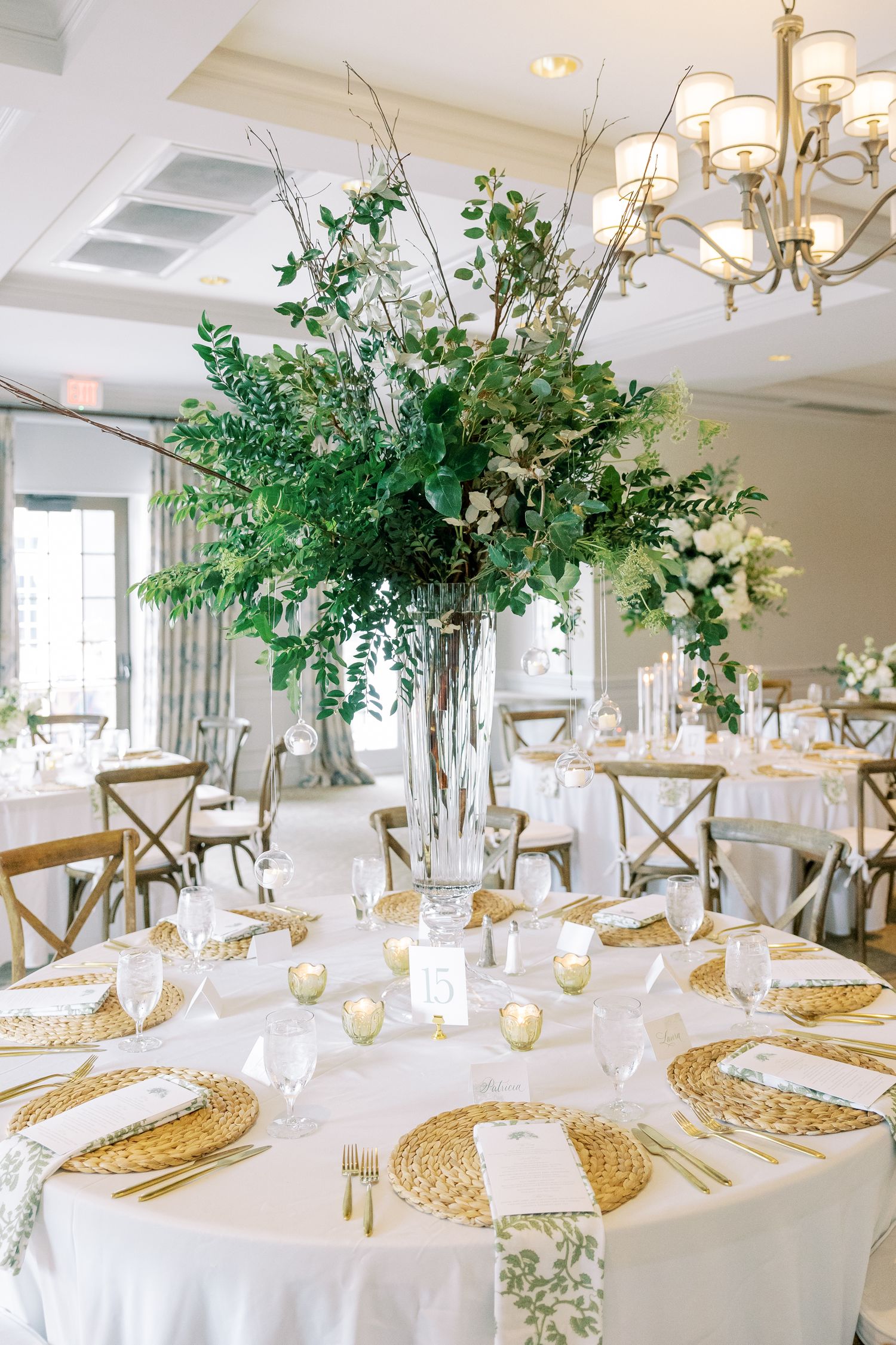 Elegant wedding reception table with gold charger plates and tall green floral centerpiece in glass vase beneath crystal chandelier.
