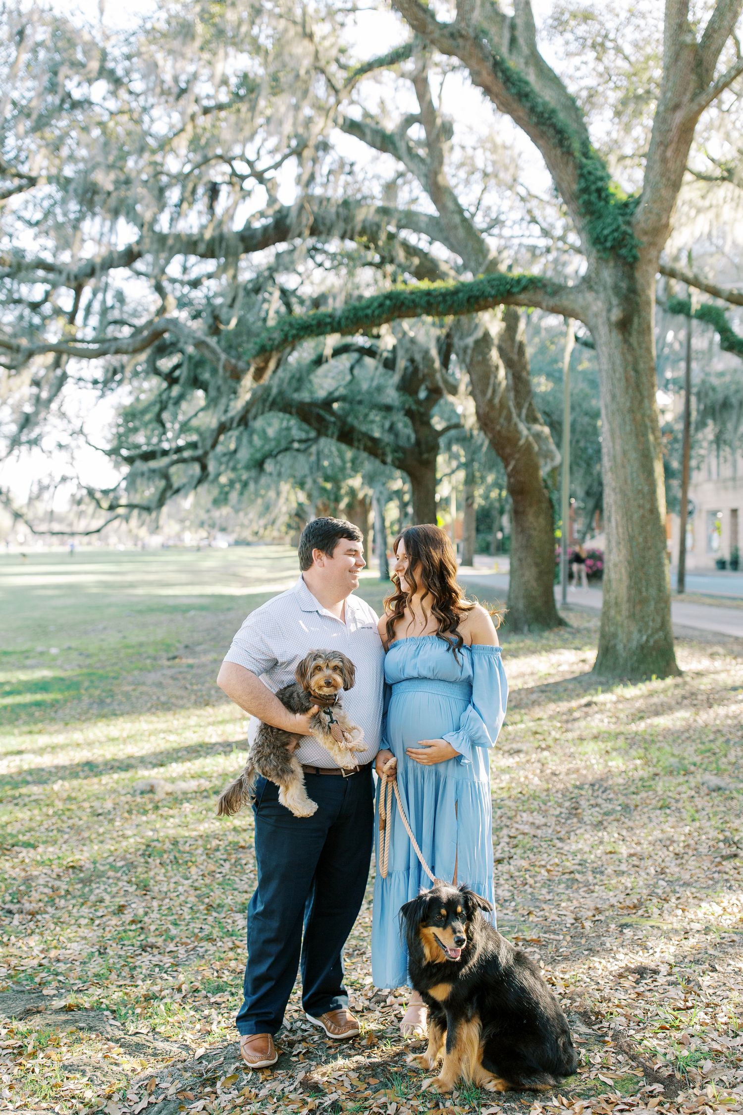 A couple poses with their dogs under moss-draped oak trees in a park setting.