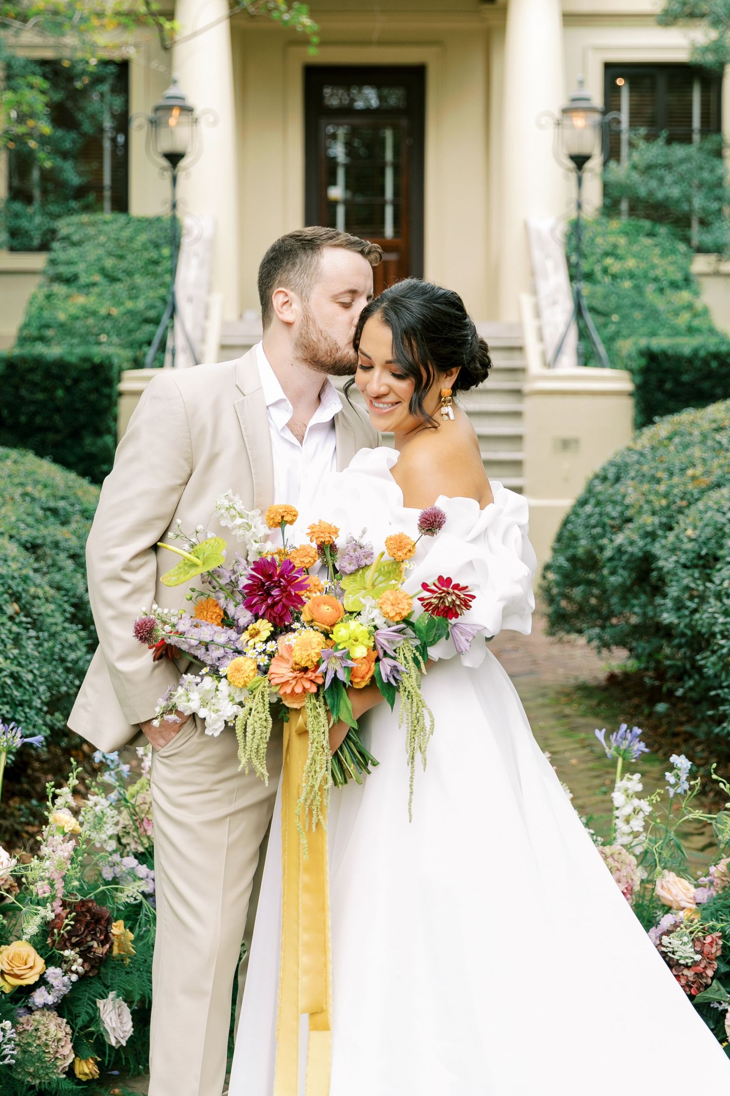 Wedding couple shares intimate moment with vibrant bouquet in front of elegant mansion entrance.