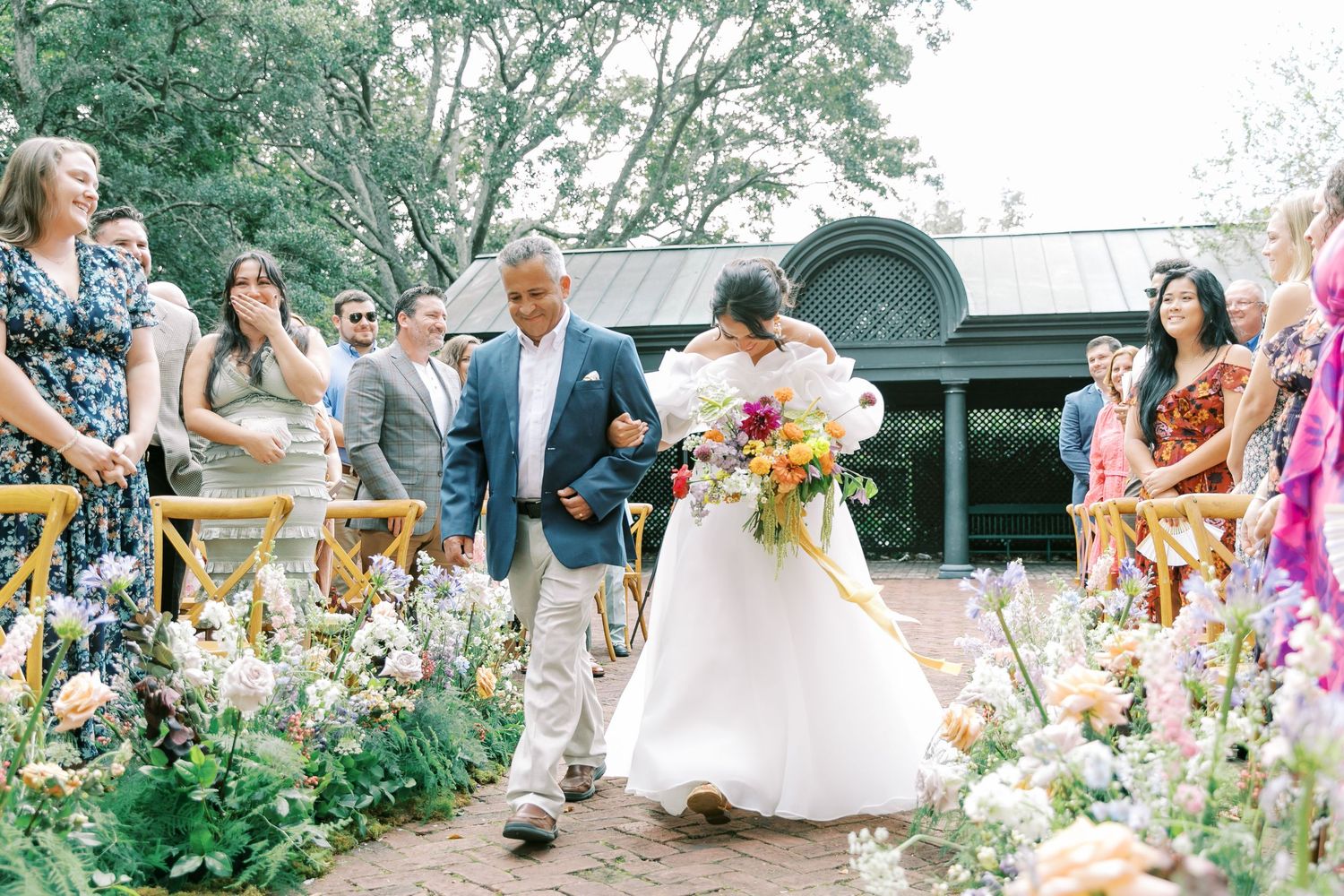 Wedding ceremony processional in a lush garden with guests watching from wooden chairs.