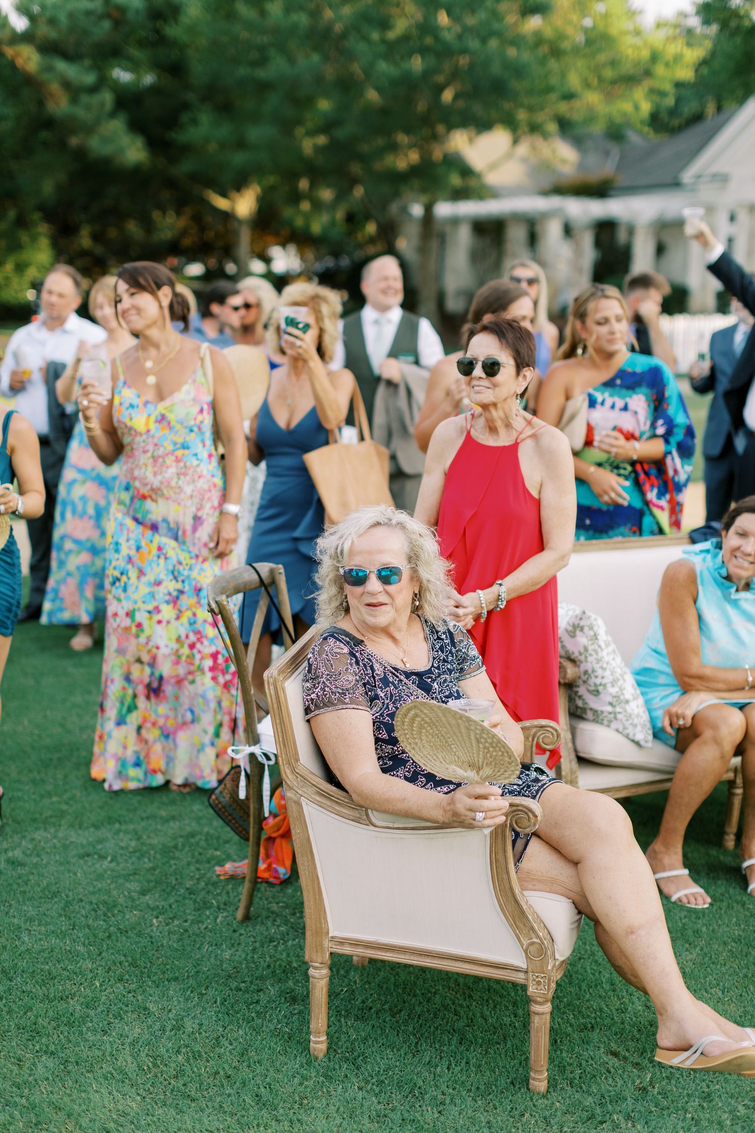 Wedding guests in colorful summer dresses gather on a lawn with a vintage chair at an outdoor celebration.