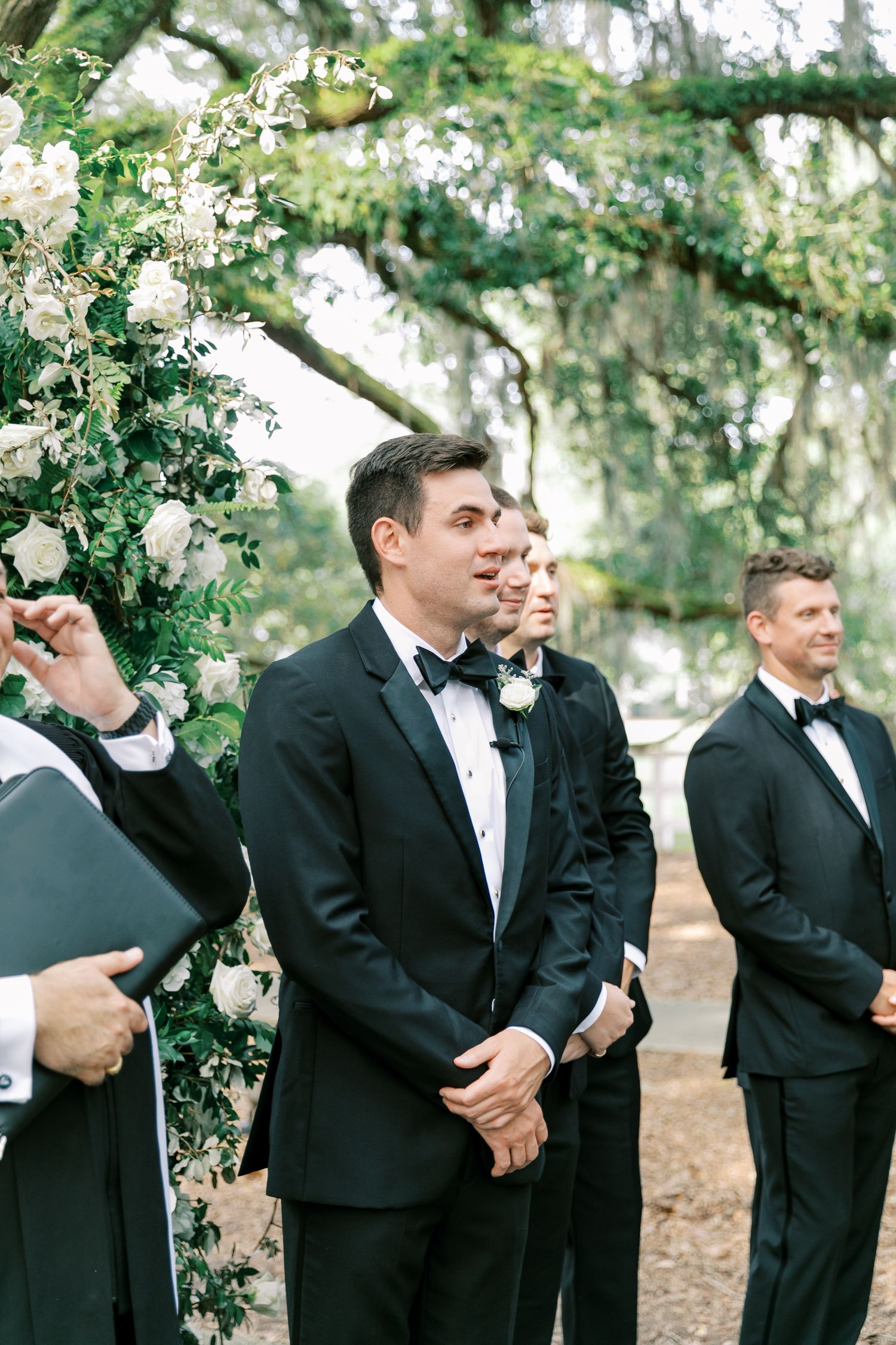Groom and groomsmen in black tuxedos standing under flowering trees at an outdoor ceremony.