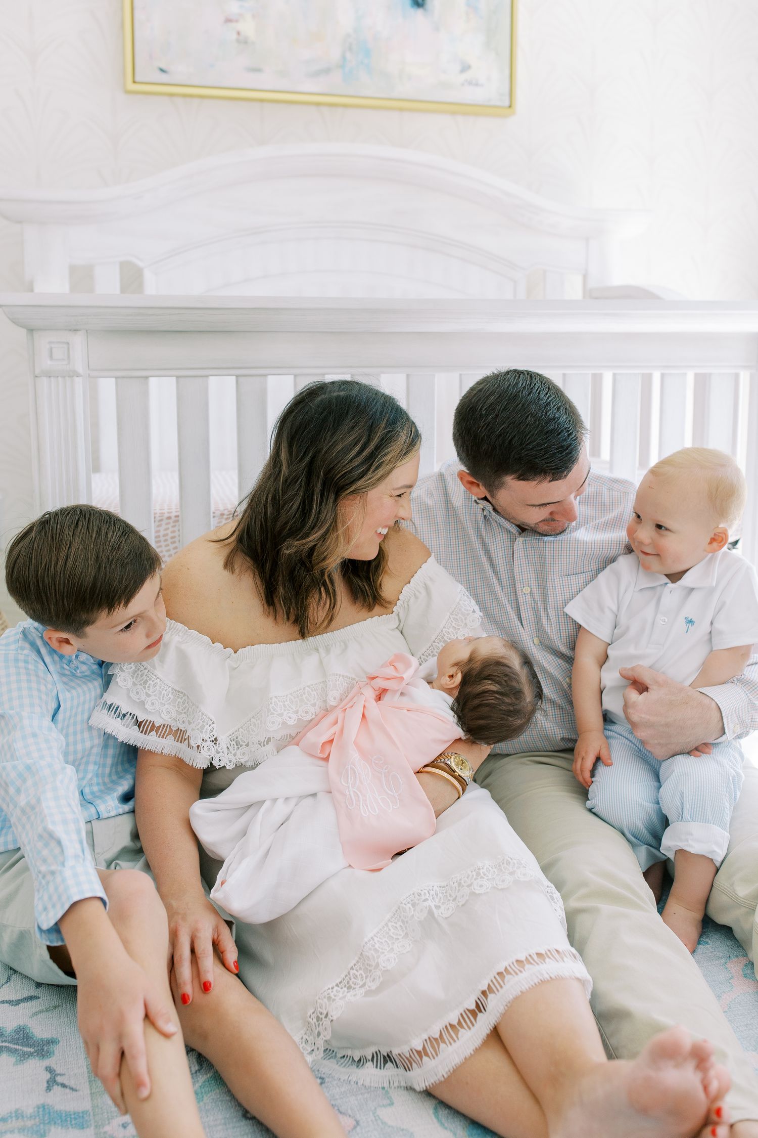 A family sits together on a white bed wearing light colored clothing while sharing an intimate moment.