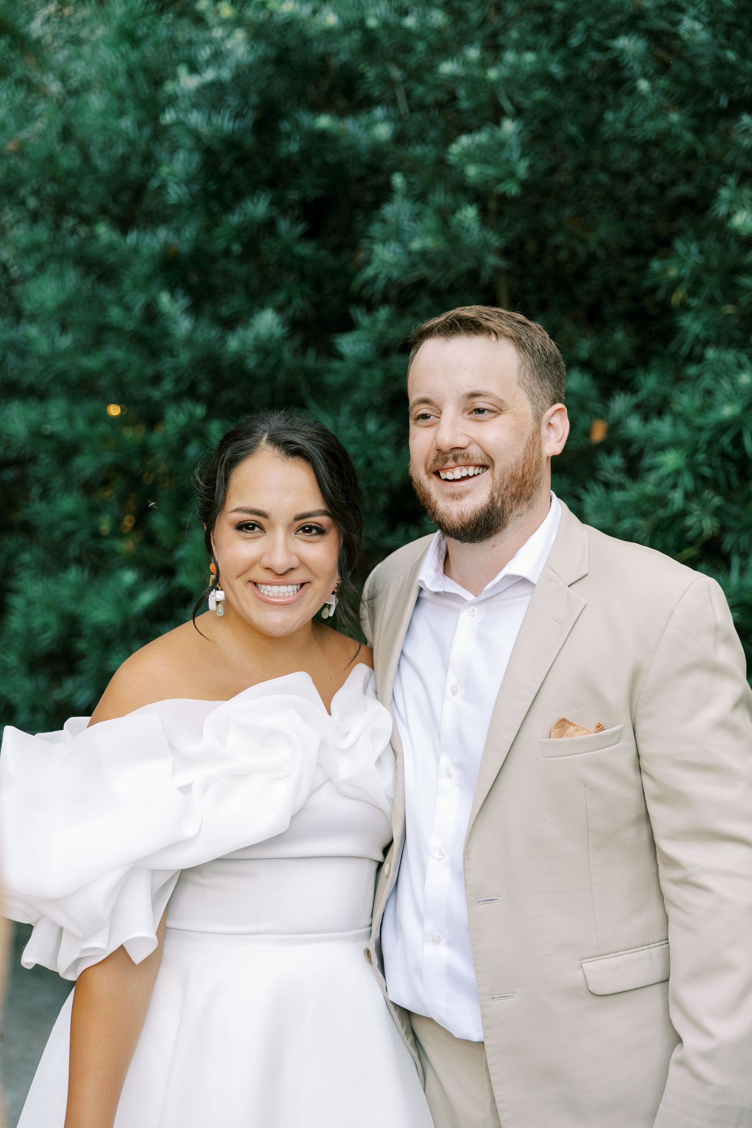 A joyful outdoor wedding portrait featuring a modern white bridal gown and light beige suit.