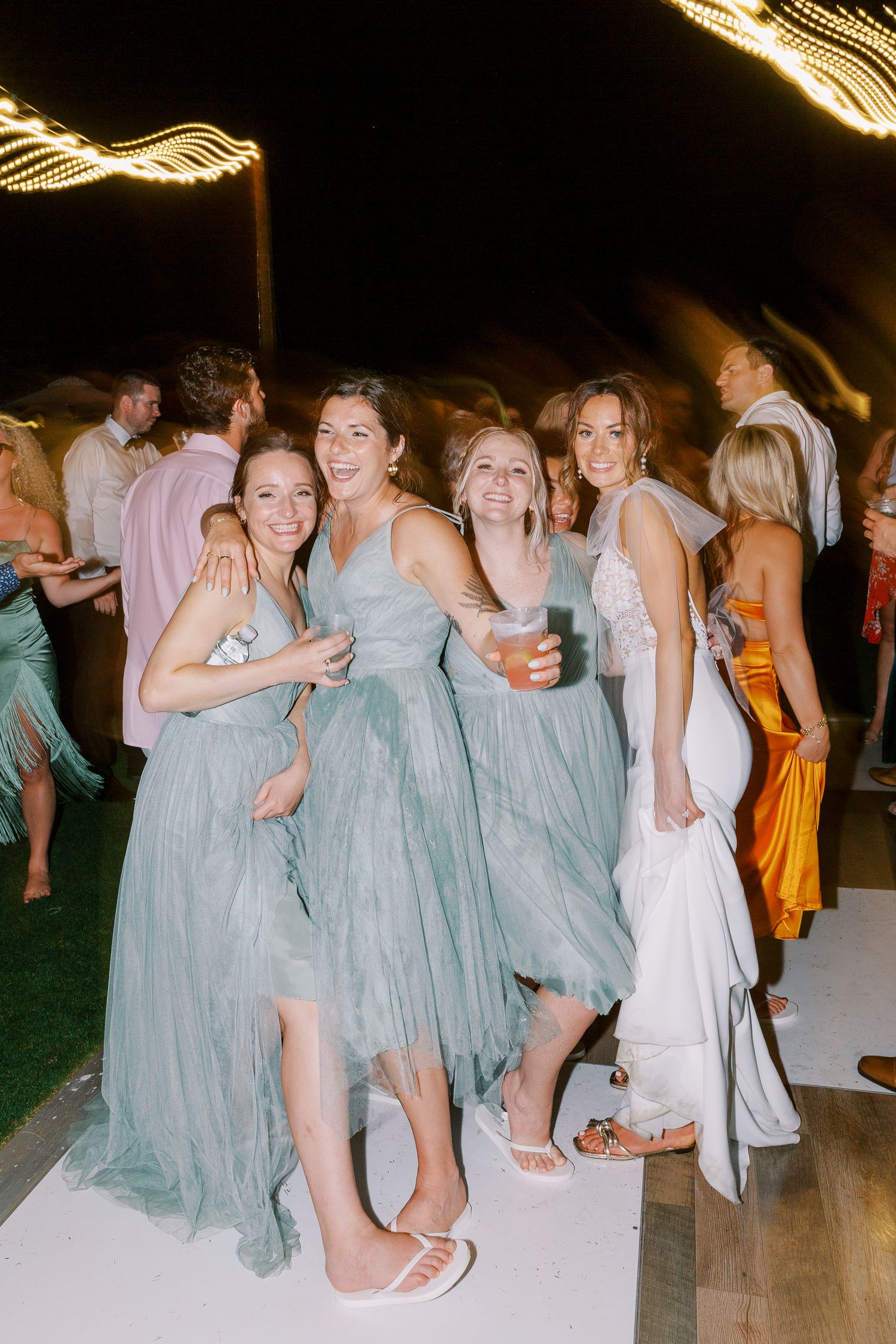 Bridesmaids in flowing dusty blue dresses laugh and dance together at a nighttime wedding reception.
