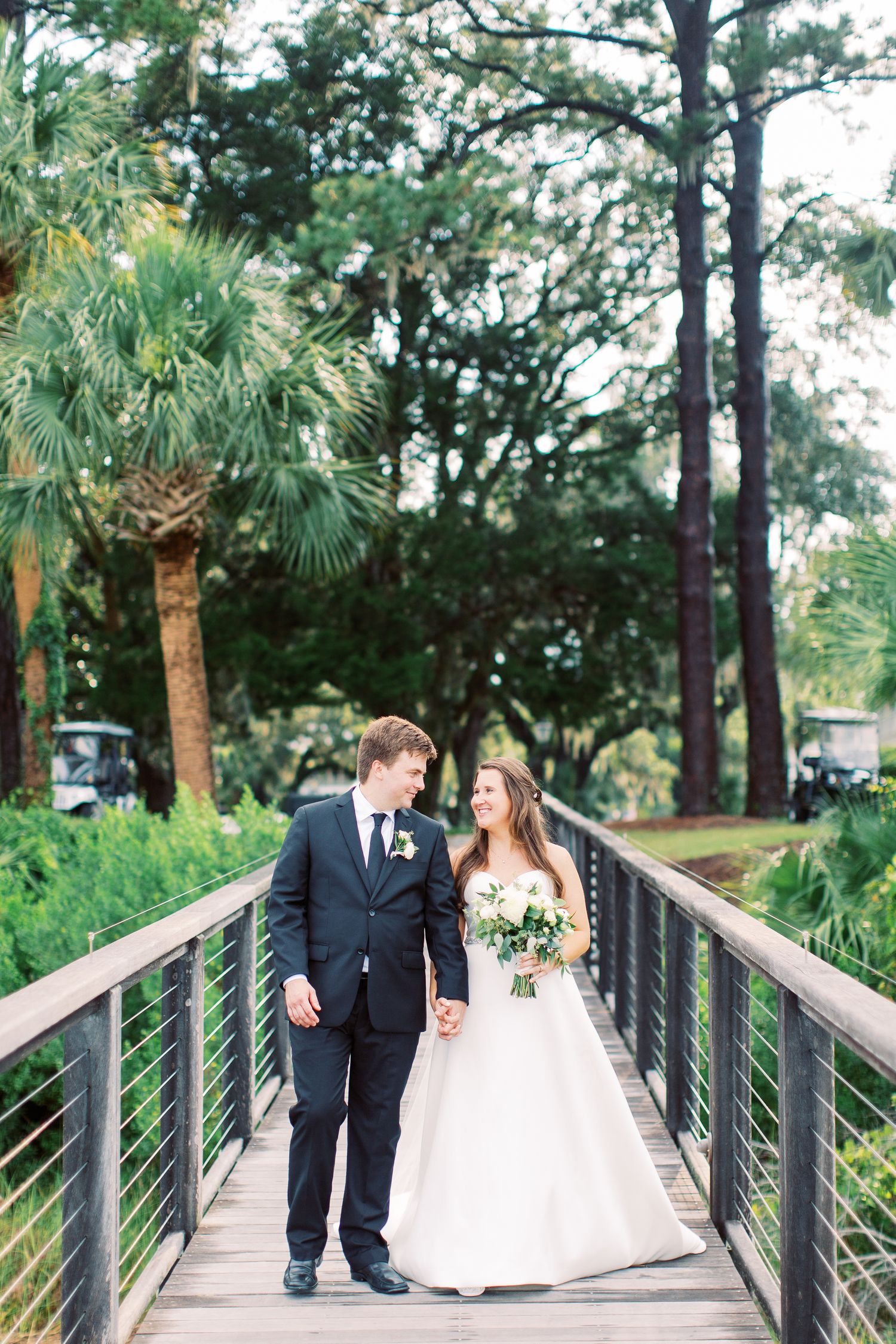 Wedding couple holds hands while standing on wooden bridge surrounded by palm trees and pine trees in outdoor setting.