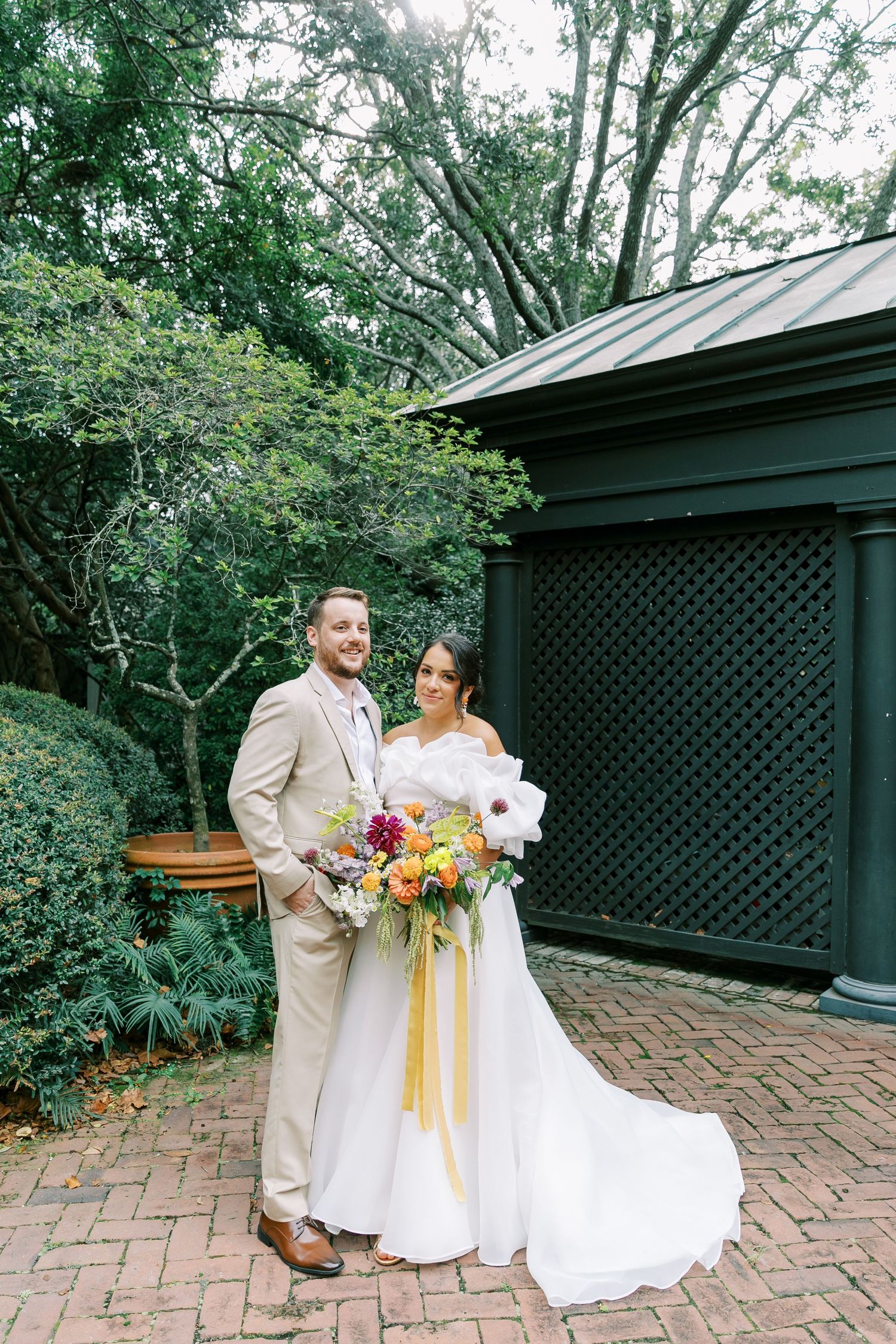 A couple in wedding attire pose together on a brick patio beside a dark green latticed structure.