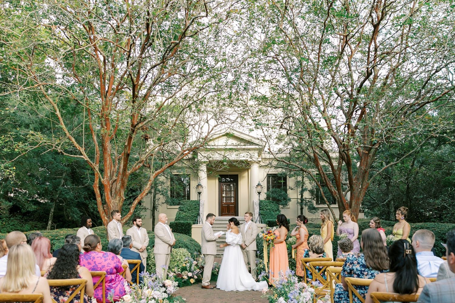 Outdoor garden wedding ceremony with guests seated beneath large trees in front of a white colonial-style building.