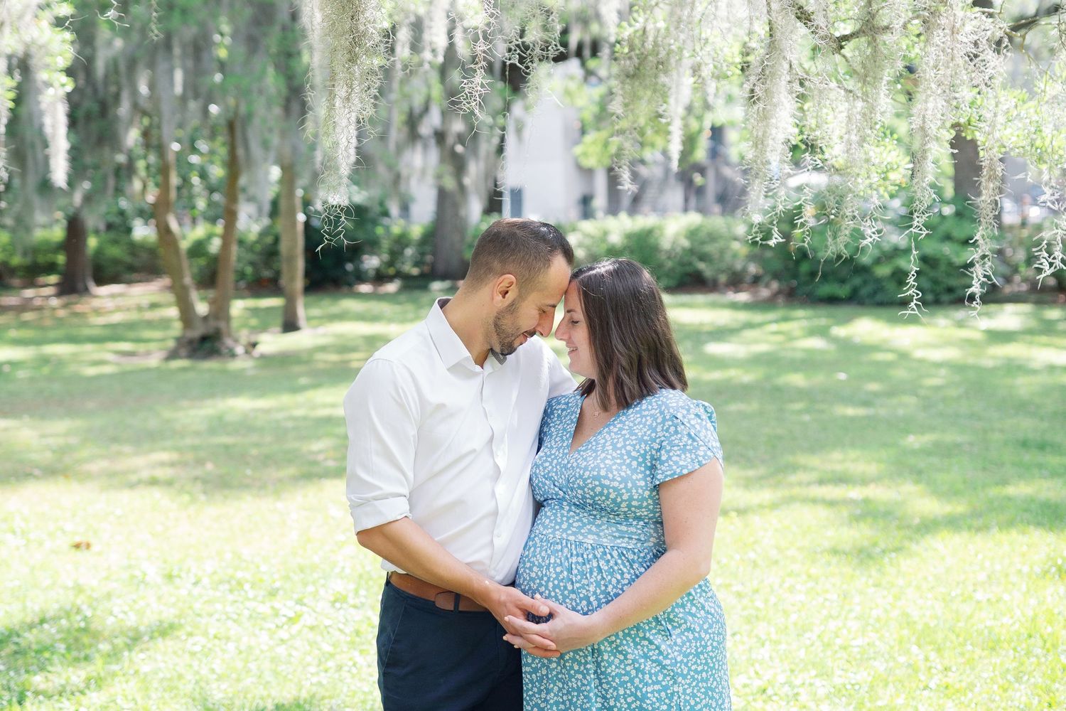 A couple shares a tender moment in a sunlit garden with Spanish moss draped trees in the background.