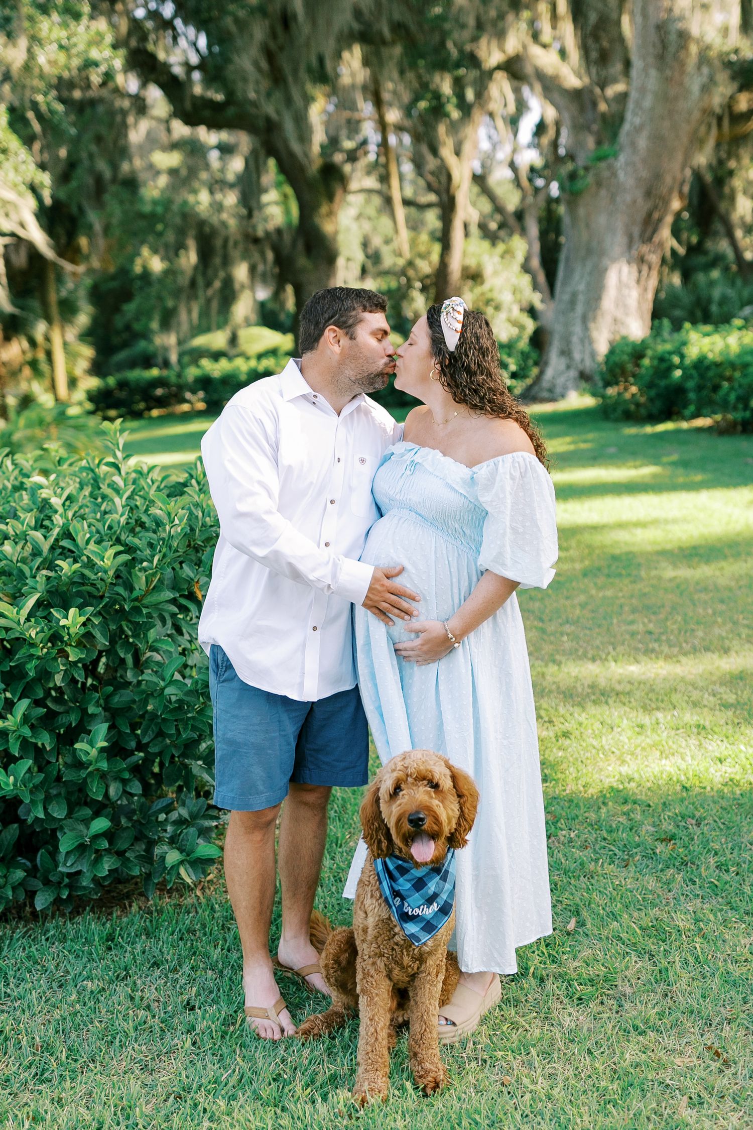 A couple in light summer attire shares romantic moments in a garden setting with their goldendoodle dog wearing a blue bandana.