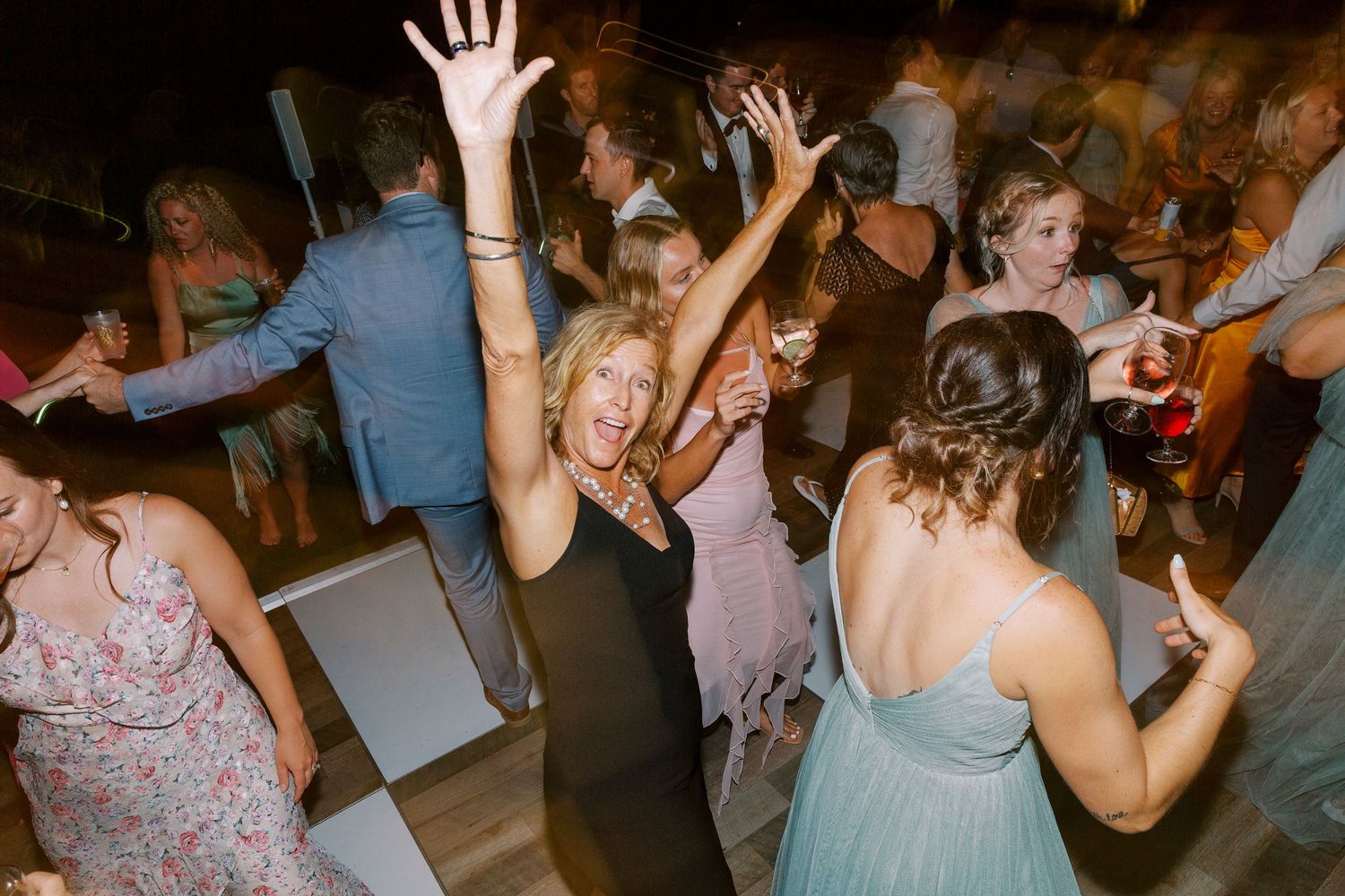 Wedding guests dance enthusiastically on a dark dance floor at a reception party.