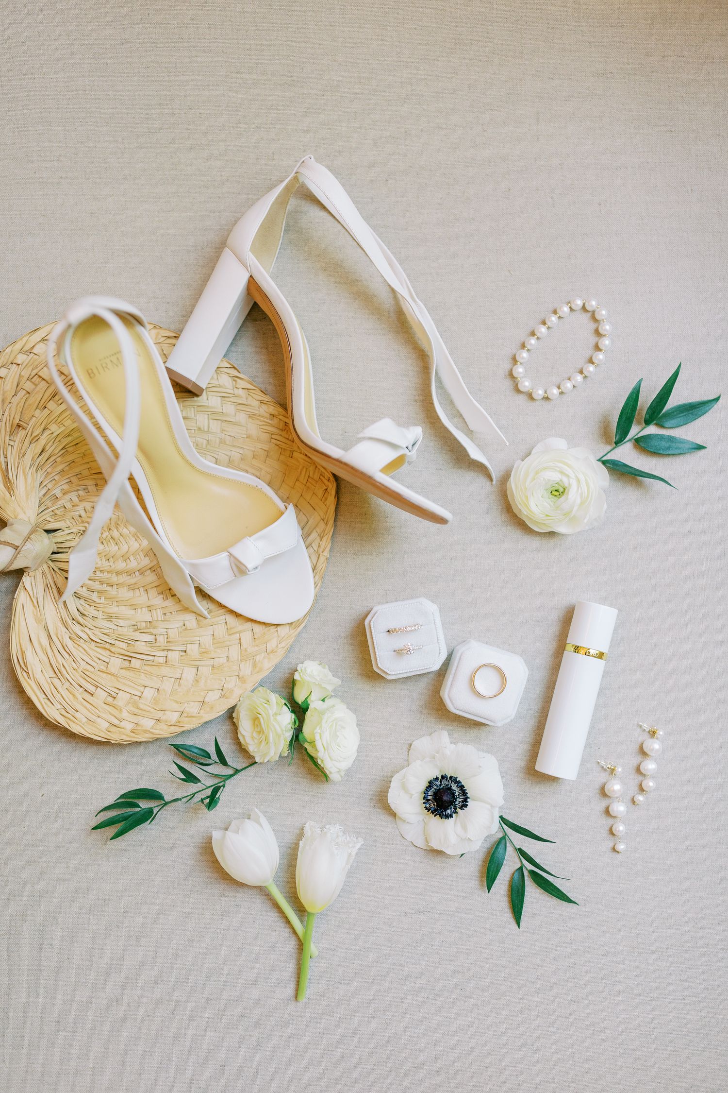 White bridal heels arranged with anemone flowers, pearl accessories, and straw fan on beige background for wedding styling.
