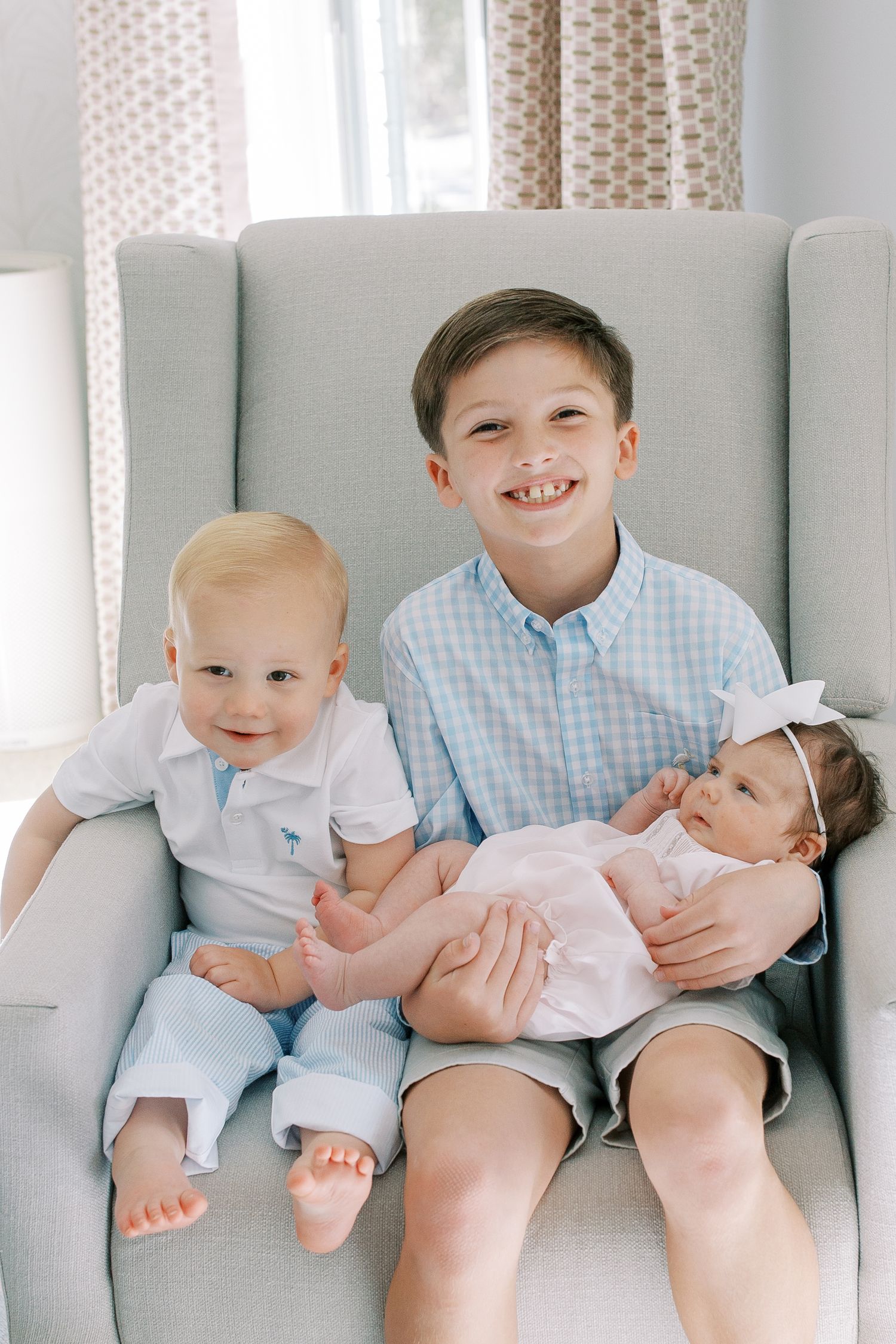 Three siblings sitting together in a cozy gray armchair by a window with polka dot curtains.