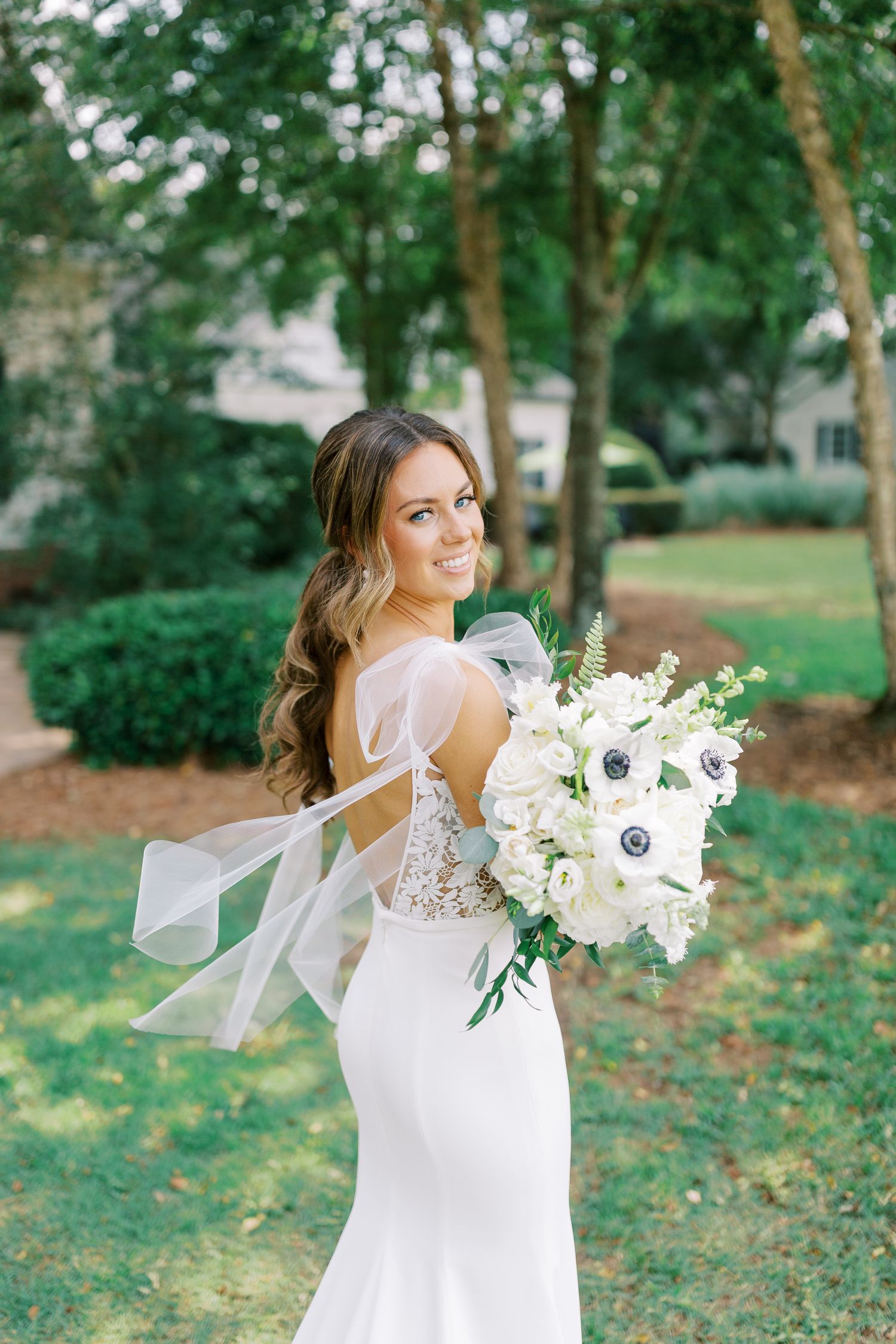Elegant bride in fitted lace wedding gown holds white and green bouquet in garden setting with flowing veil and ribbons.