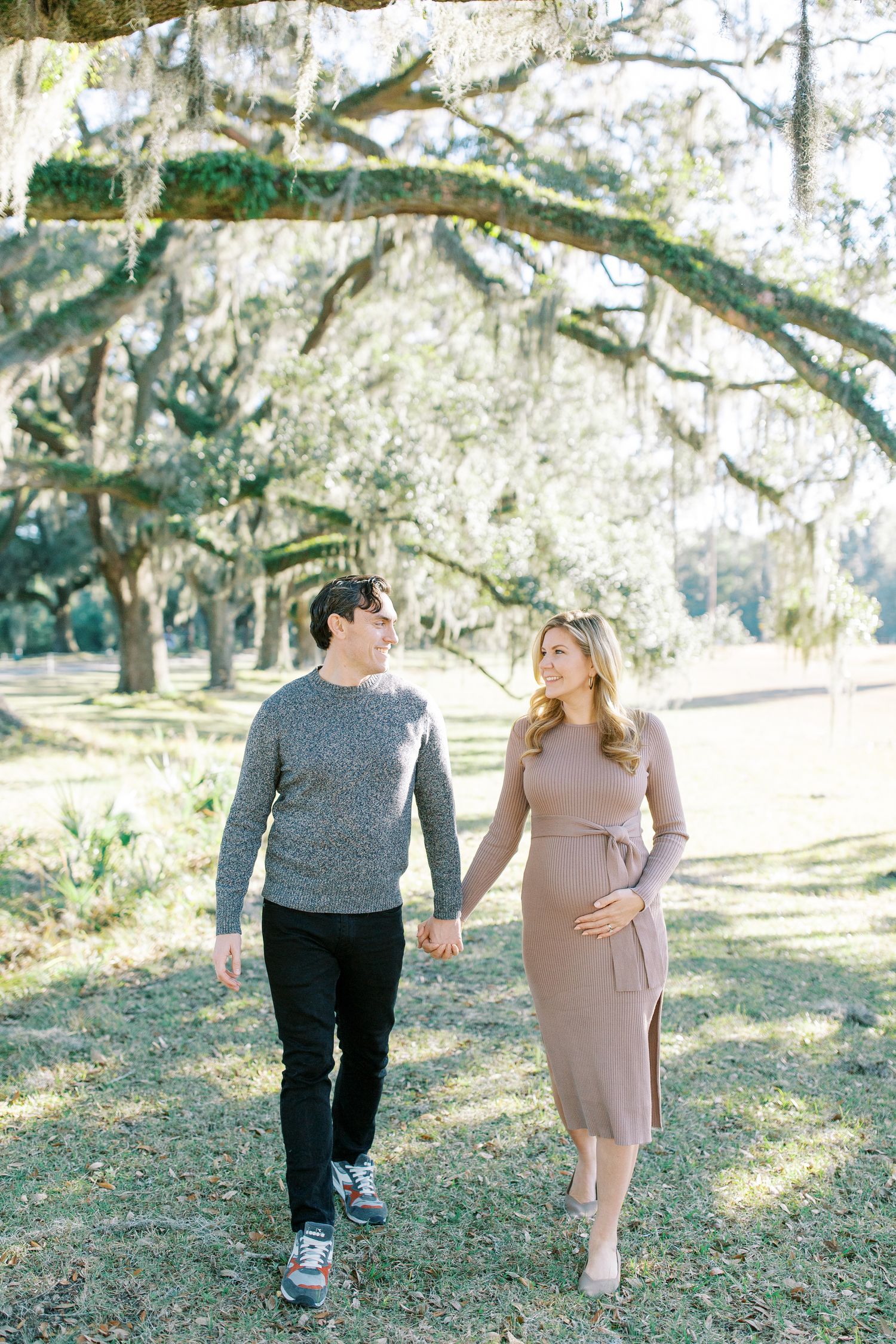 A couple walks hand in hand through a scenic park with Spanish moss draped oak trees overhead on a sunny day at wormsloe state park