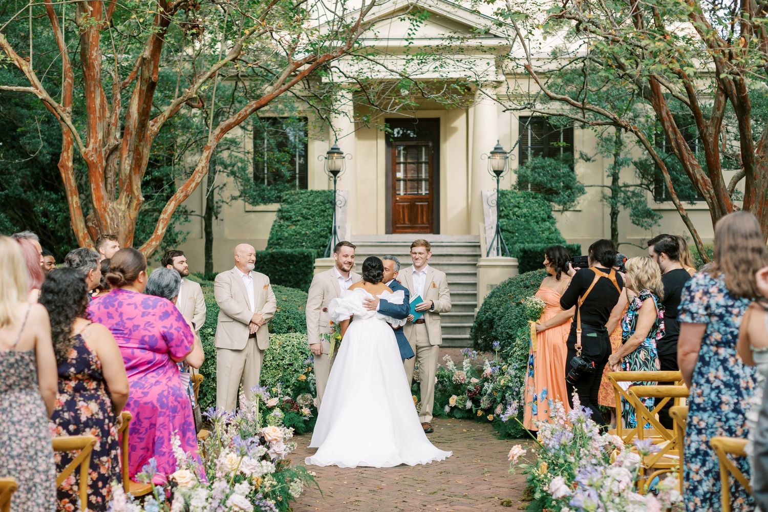 A scenic outdoor wedding ceremony setup with guests seated in chairs under tall trees.
