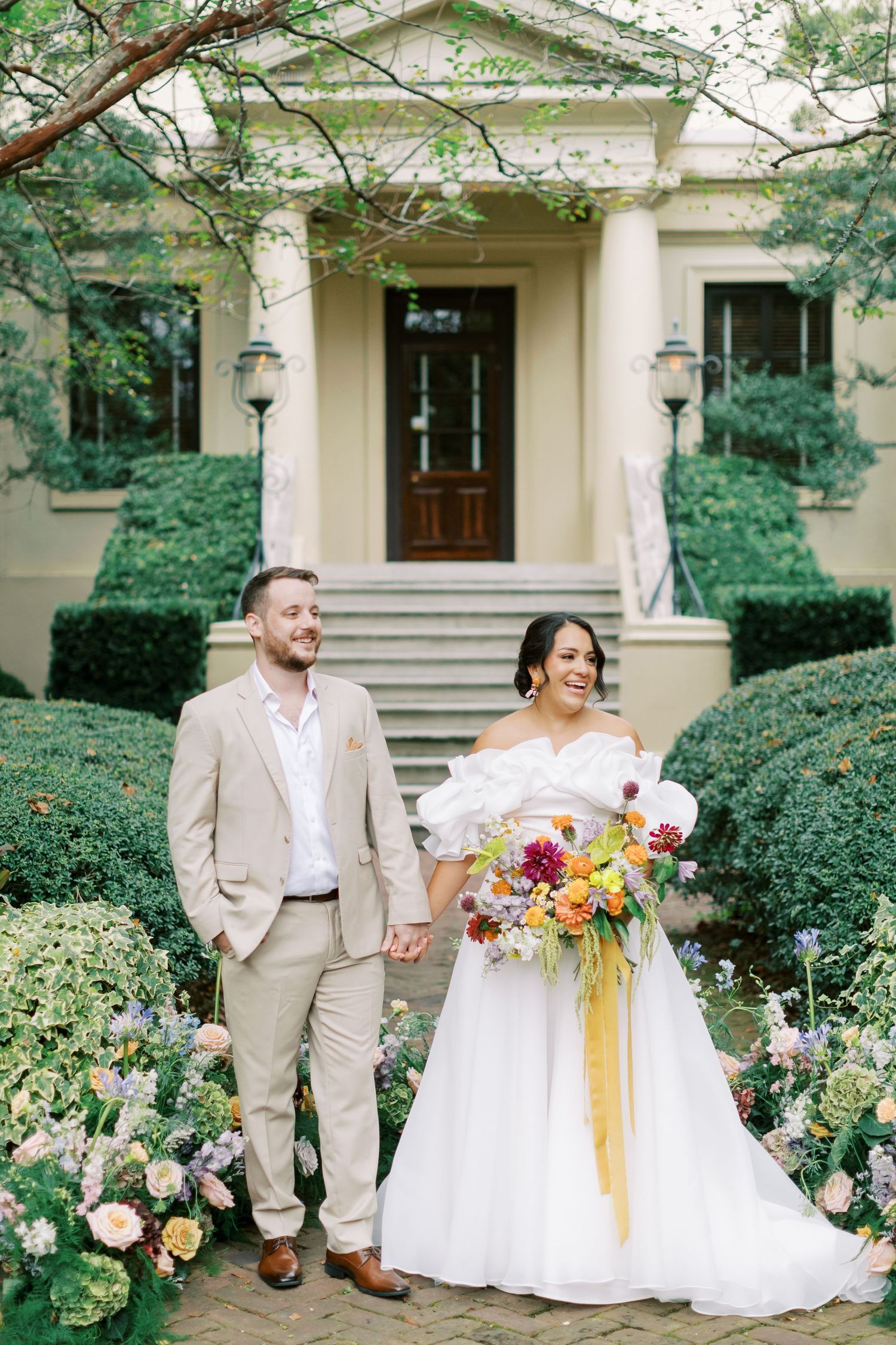 A wedding couple poses for romantic portraits in front of a stately building with manicured gardens.