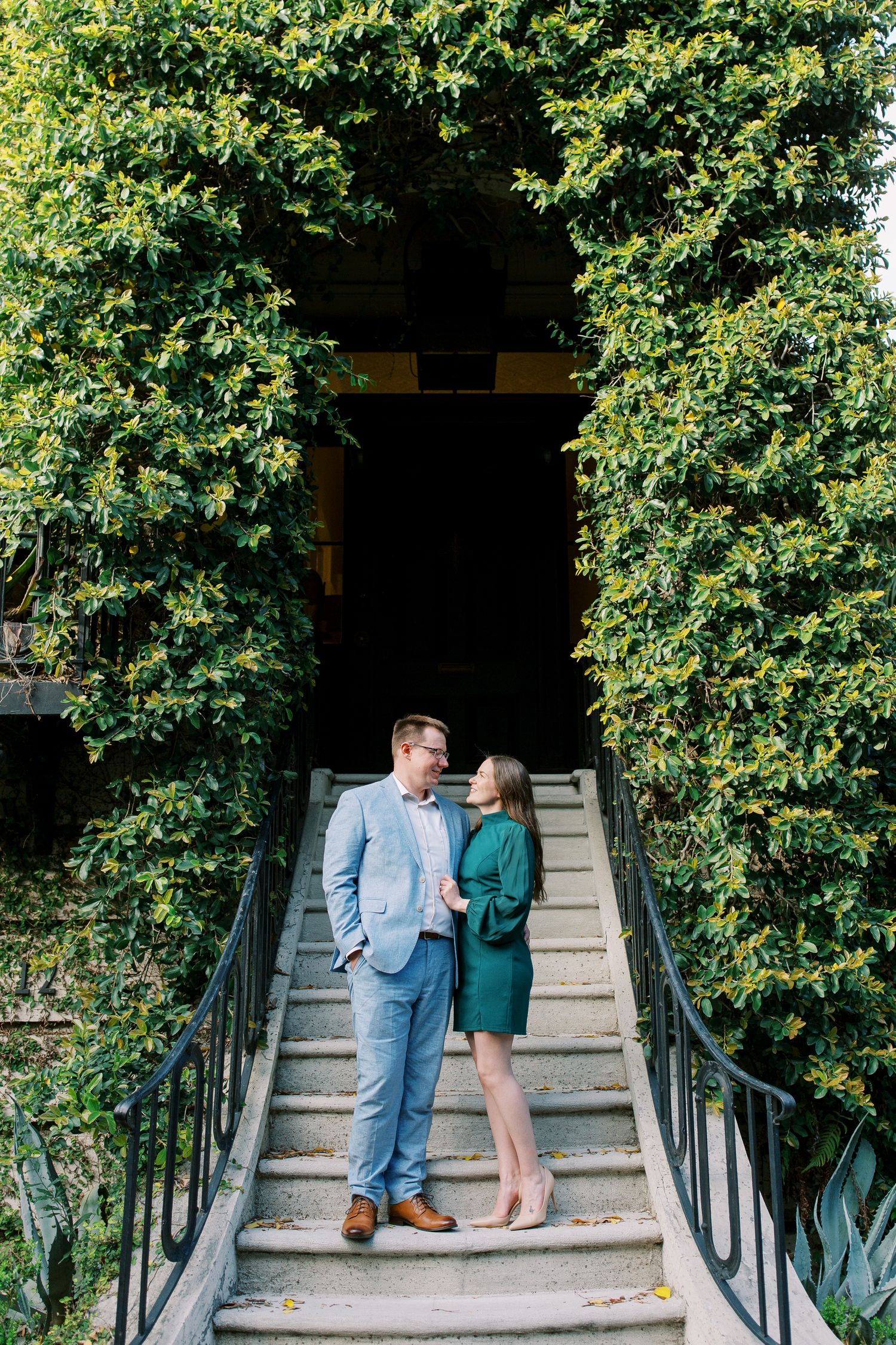 A couple stands together on stone steps framed by green foliage and iron railings.