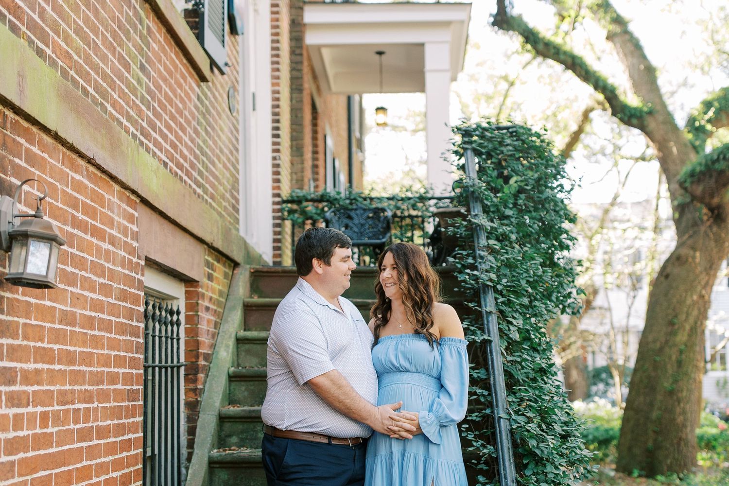 Two people in romantic pose stand beside brick wall and greenery outside a historic townhouse steps.