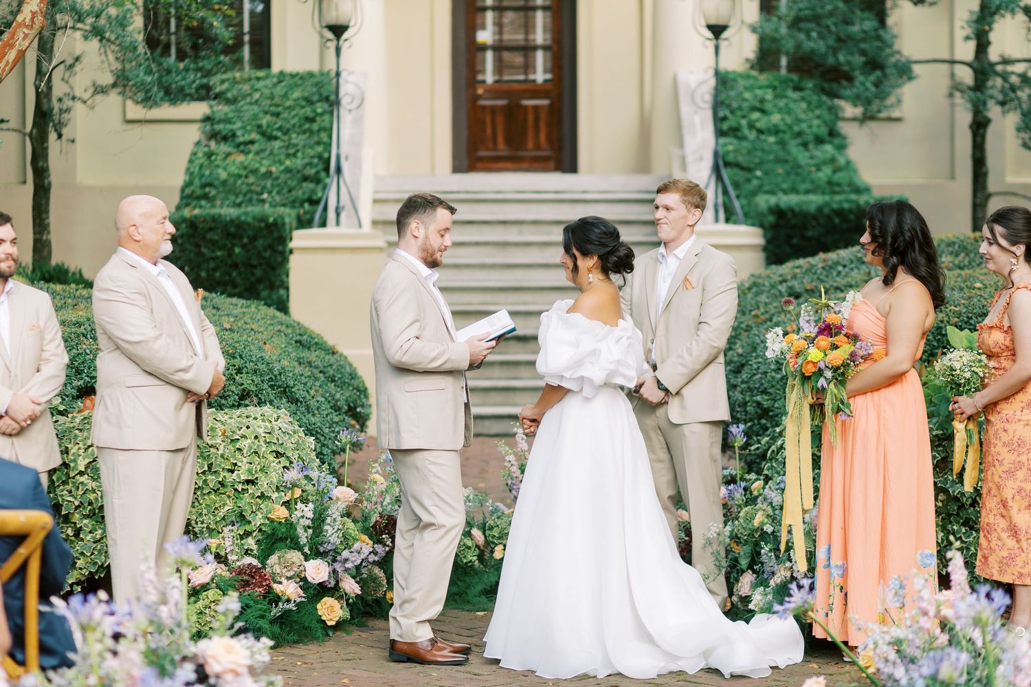 An elegant outdoor wedding ceremony setup with pink floral arrangements and cream-colored suits.