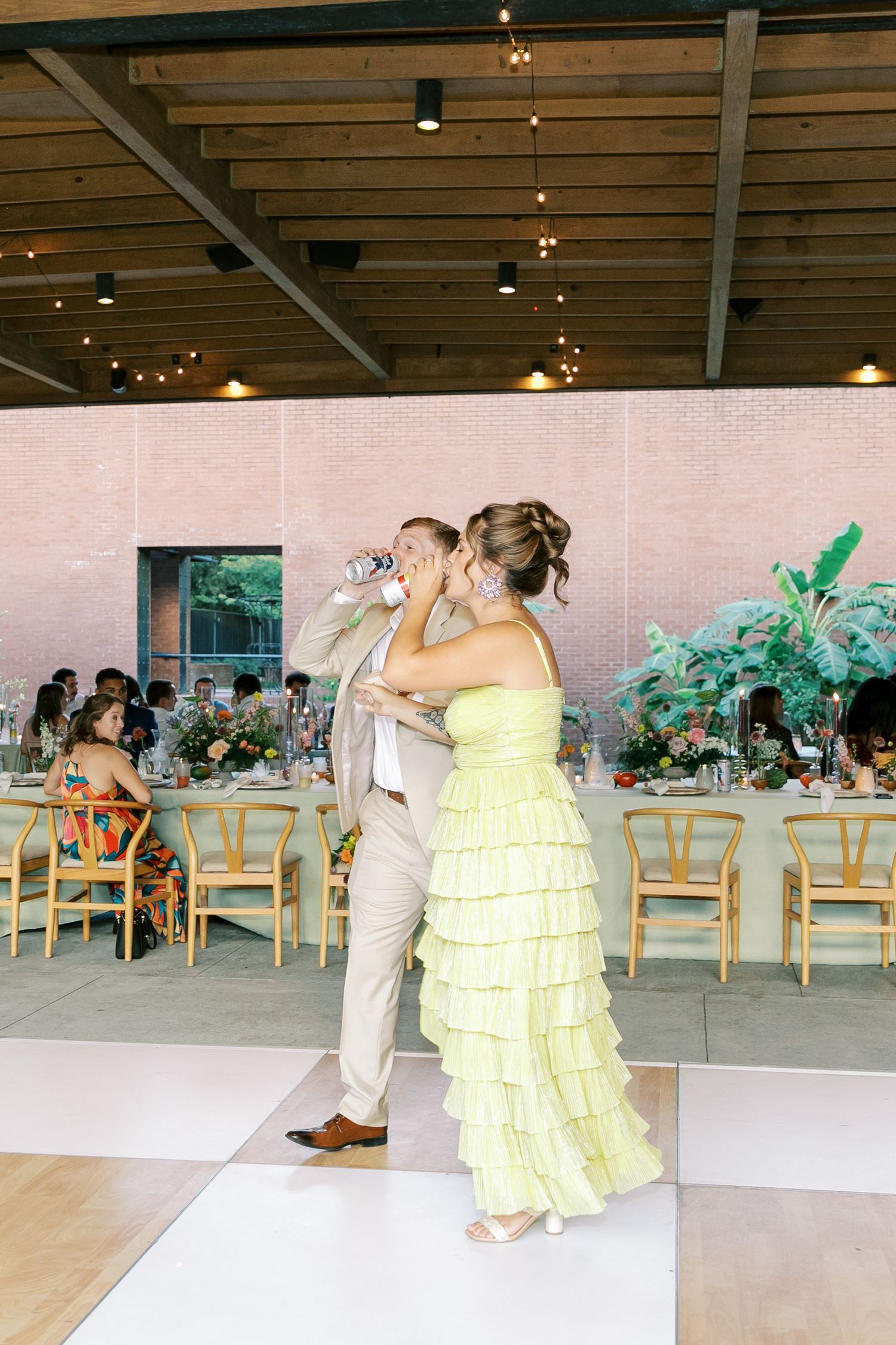 A couple dancing in formal attire with a yellow ruffled dress under string lights at a reception.