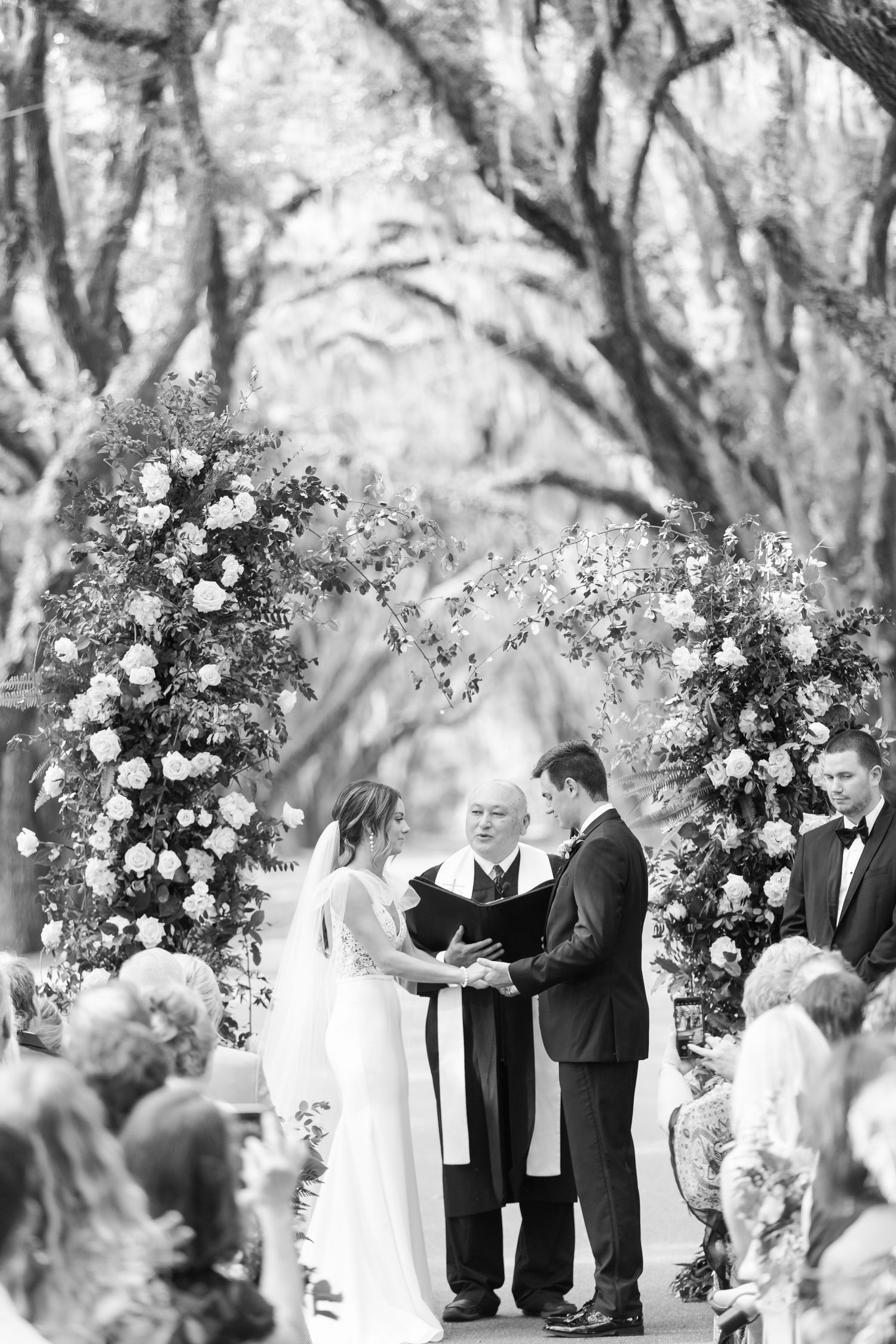 A romantic outdoor wedding ceremony under oak trees with a floral arch and gathered guests.