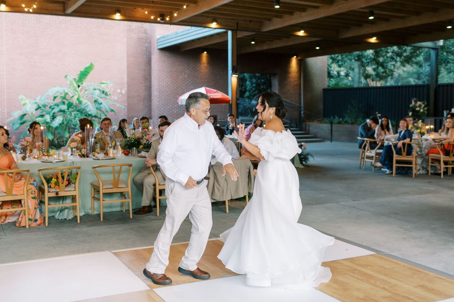 A couple shares their first dance on a wooden dance floor during their outdoor reception at a modern event space.