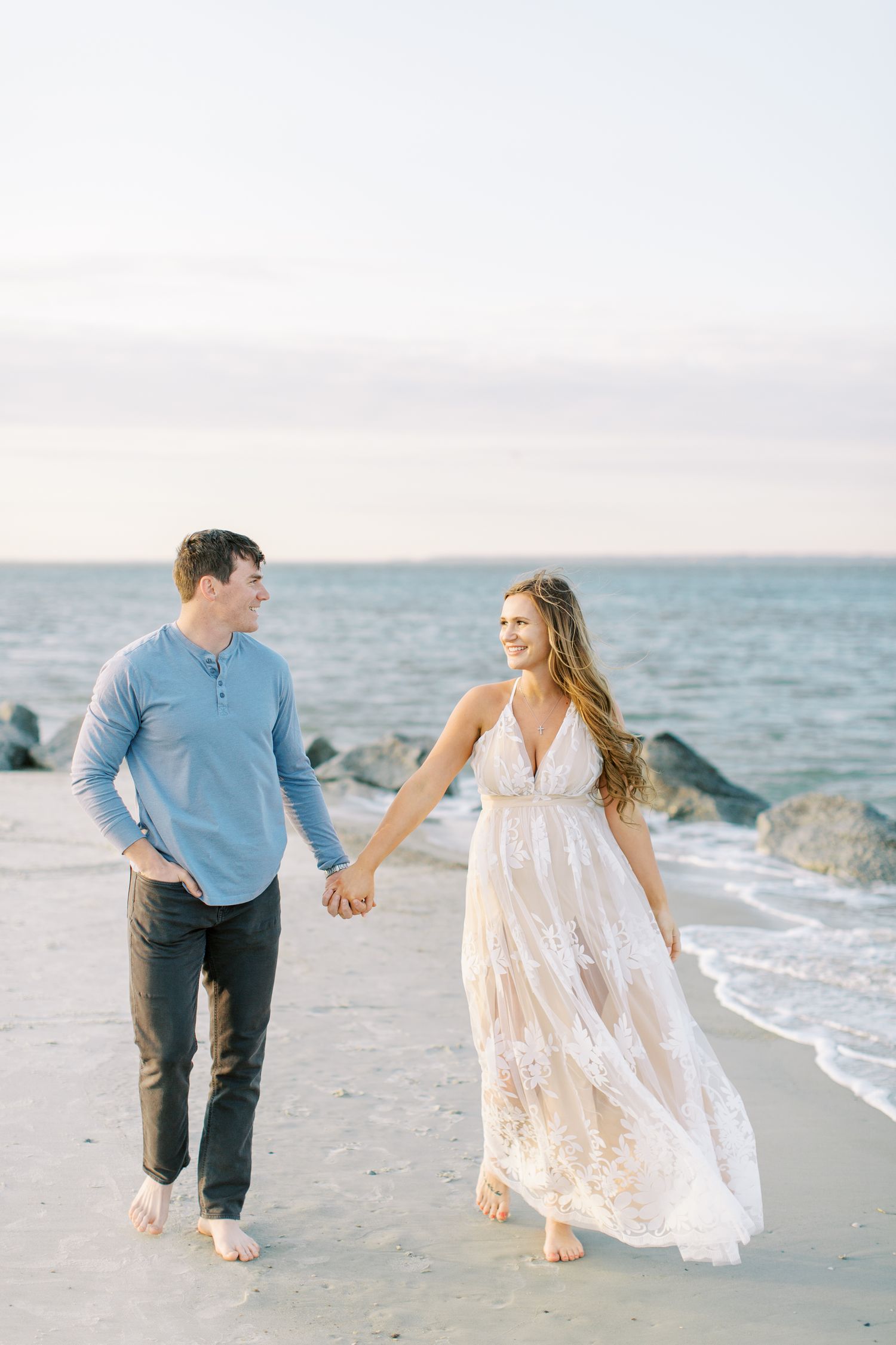 A couple walks barefoot along a sandy beach at sunset holding hands, with waves and rocks in the background.